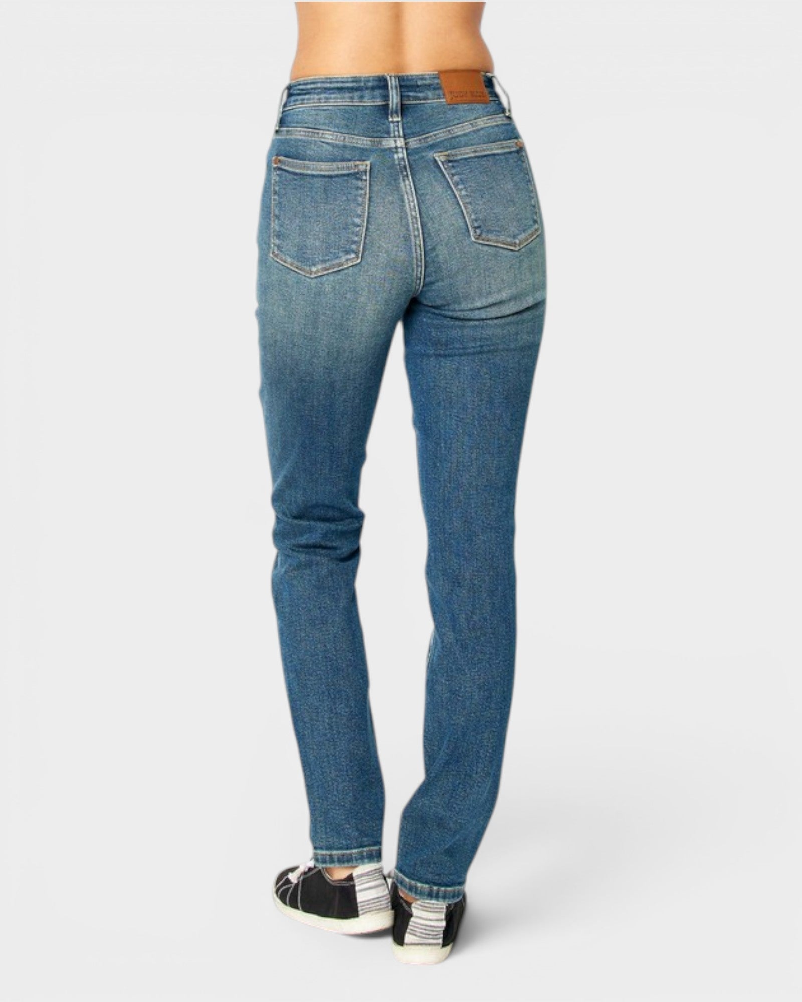 Rear view of a person wearing medium-wash blue denim jeans with black and white sneakers, shown against a light gray studio background.
