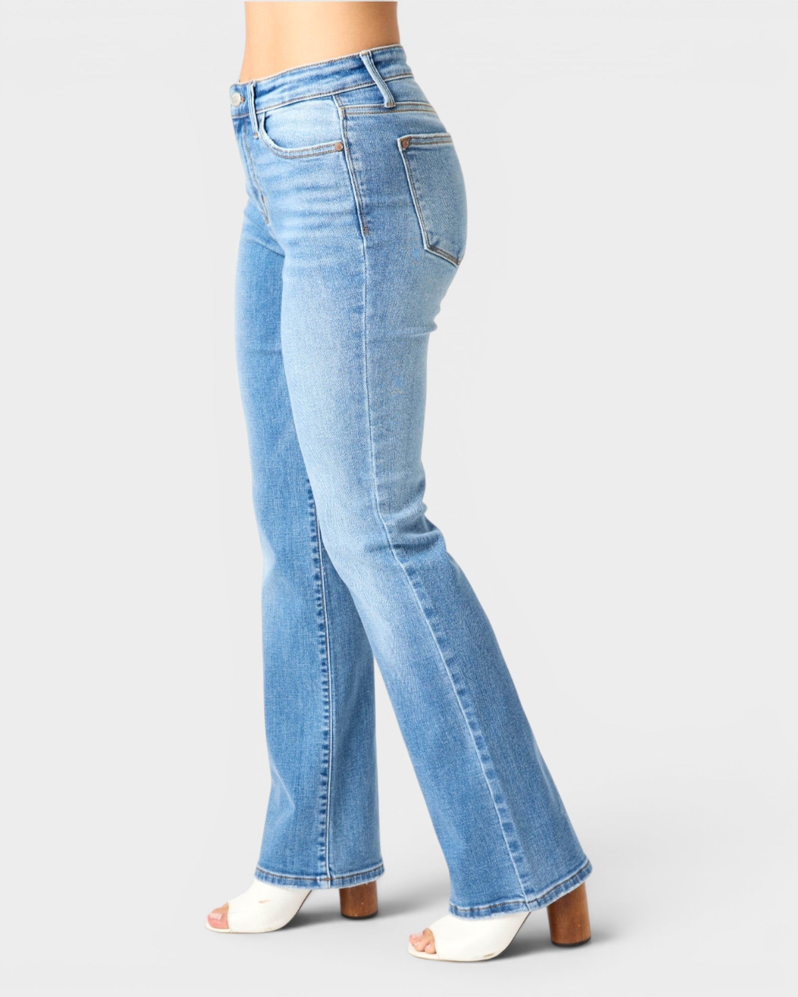 Light blue high-waisted flare jeans shown on a model from the side, paired with white heeled sandals against a light gray studio background.
