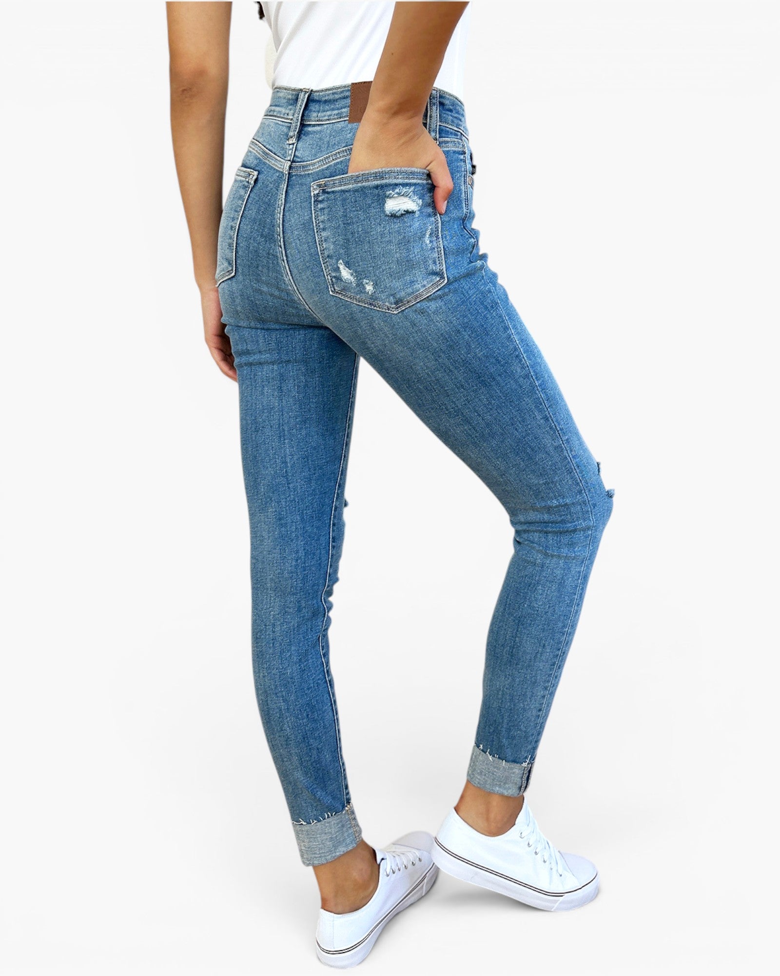 A person wearing blue high-waisted skinny jeans with distressed details and cuffed hems, paired with white sneakers, shown from a rear three-quarter angle against a plain white background.
