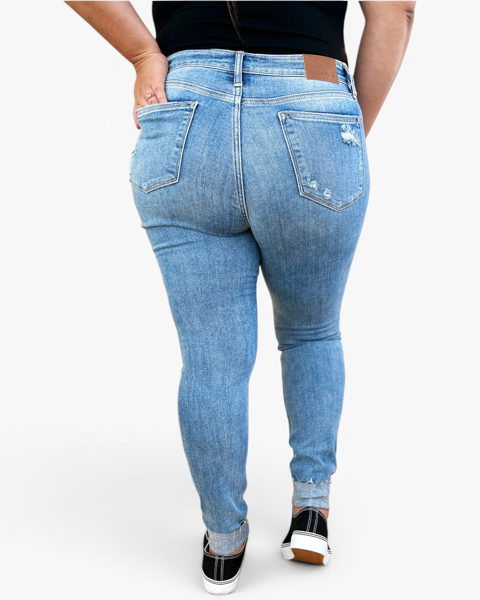 A person shown from the back wearing light-wash blue skinny jeans with distressed details on the back pockets, paired with a black top and black sneakers, against a plain white studio background.