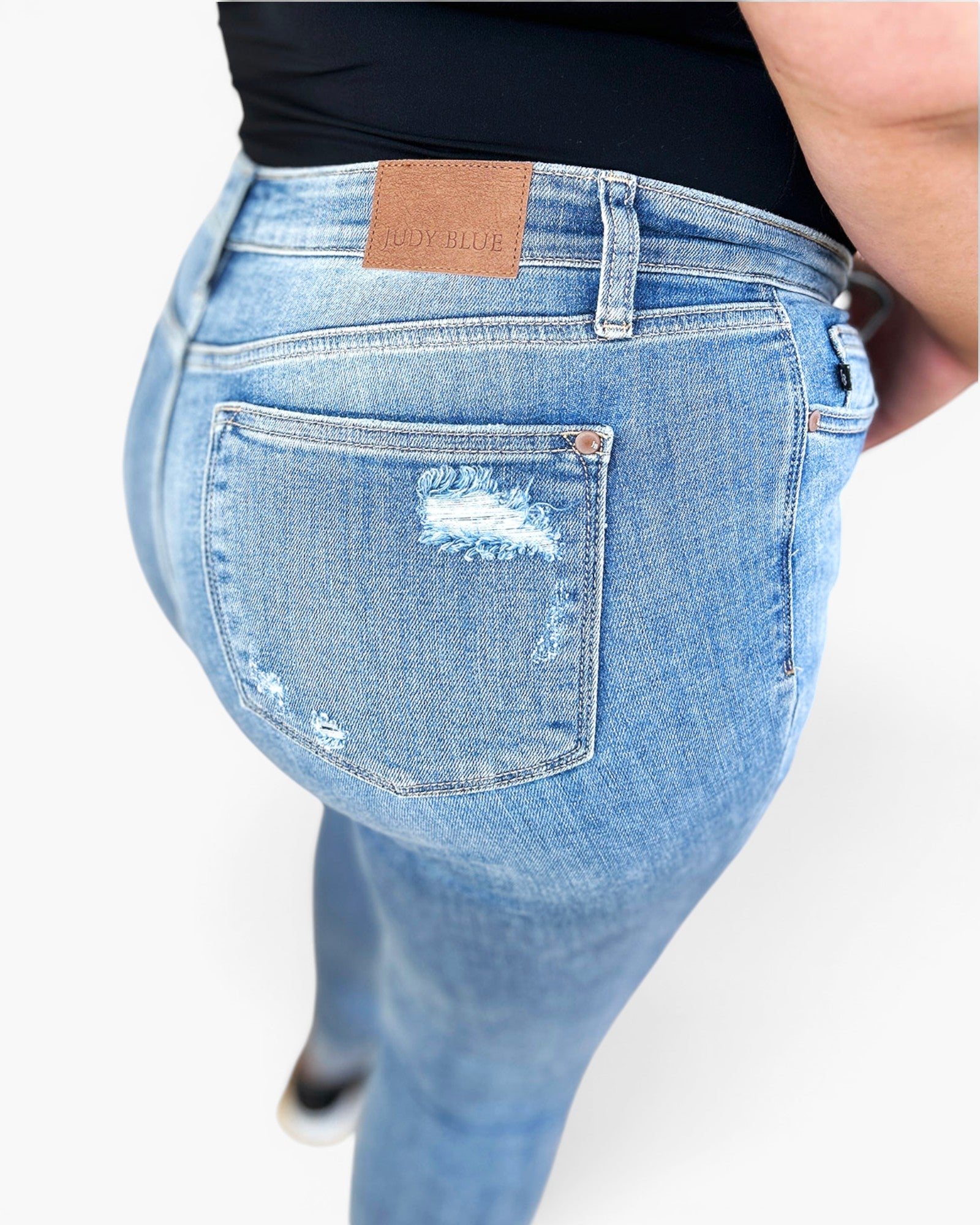 Close-up back view of a person wearing light blue distressed skinny jeans with a tan leather brand patch on the waistband, photographed against a plain light background.