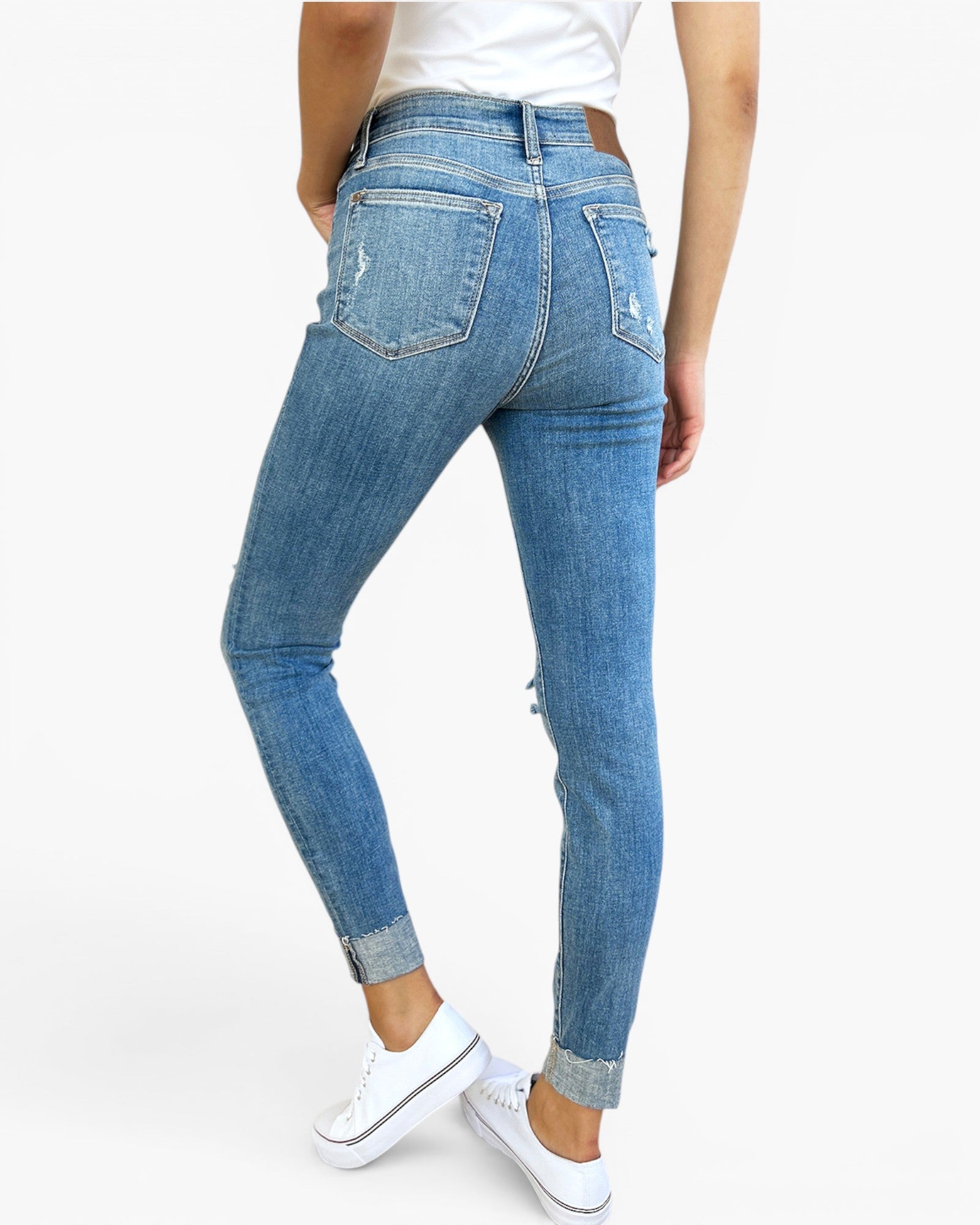 A person wearing light blue high-waisted skinny jeans with distressed details and rolled cuffs, paired with a white top and white sneakers, shown from a rear three-quarter angle against a plain white background.