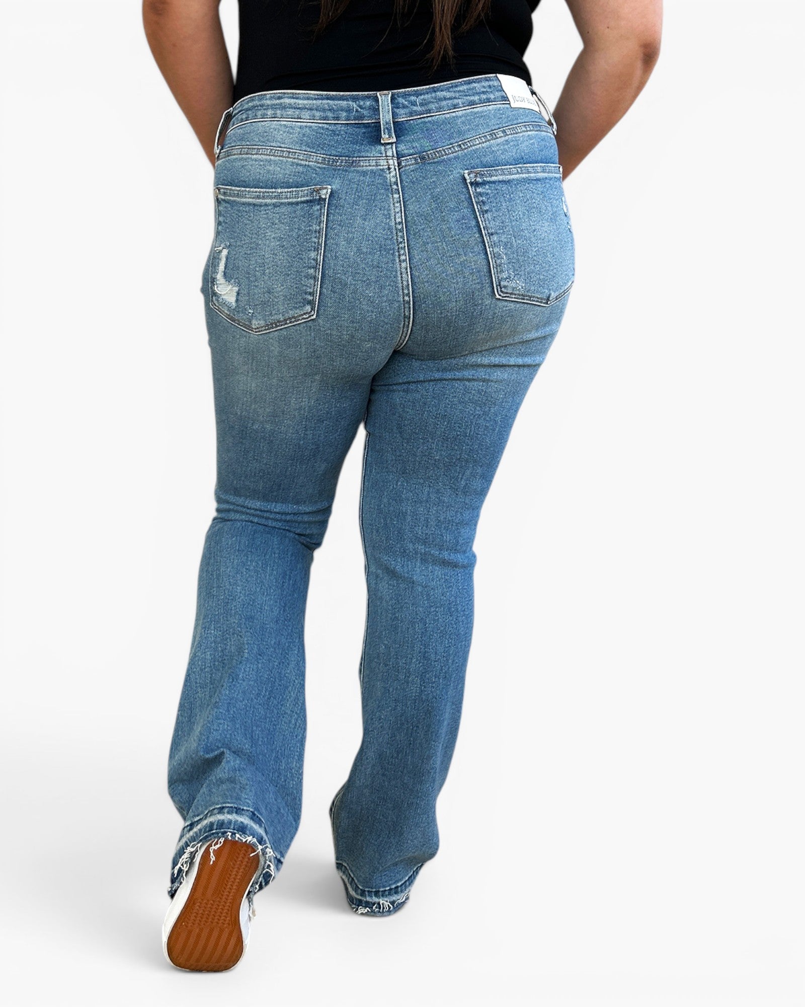 Rear view of a person wearing light blue denim jeans with a slight flare and distressed detail on the left back pocket, paired with a black top and brown shoes, against a plain white background.