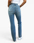 Rear view of a person wearing medium-wash blue denim jeans with a distressed patch on the left back pocket, paired with a white top and white sneakers against a light gray studio background.