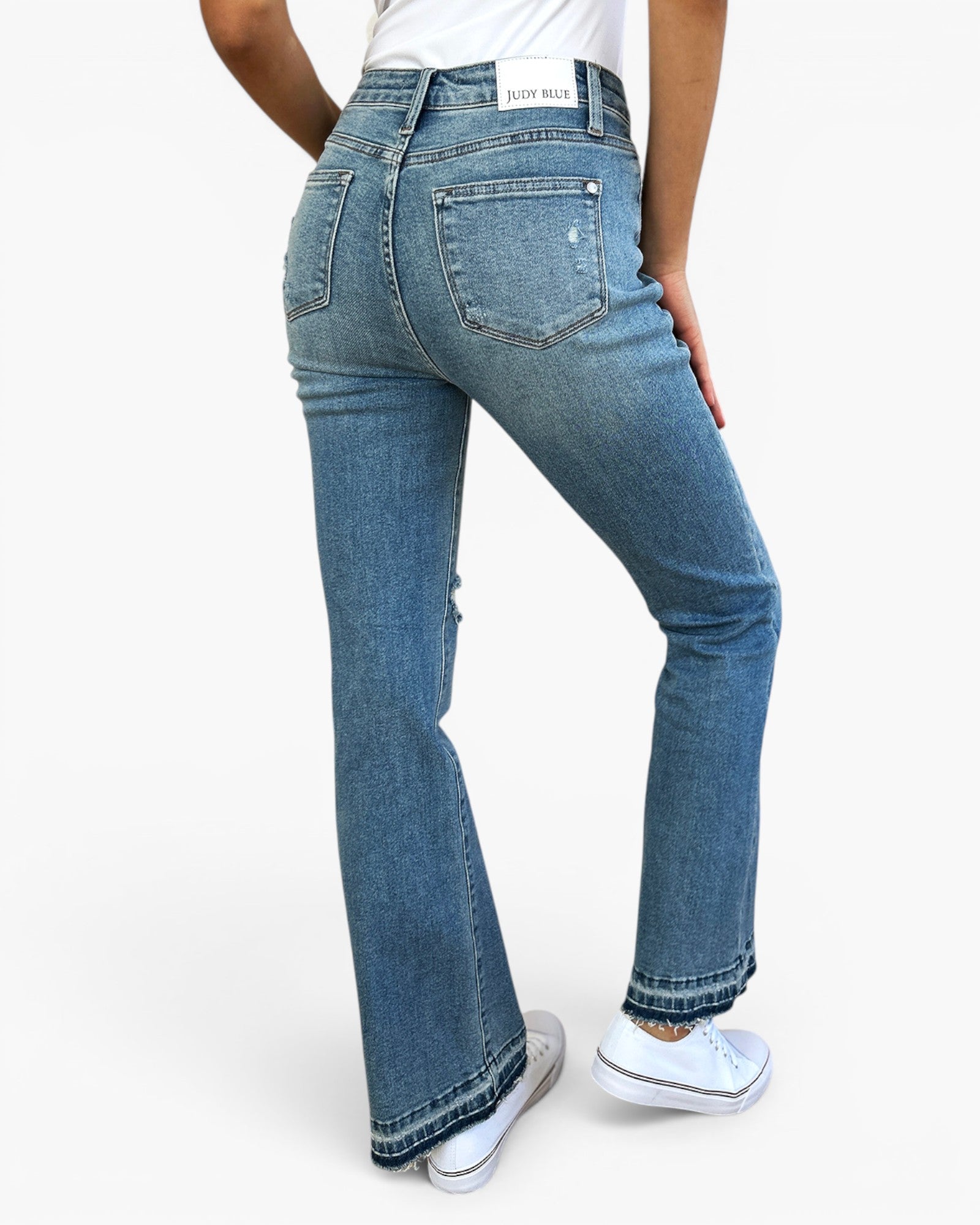 A person wearing blue denim jeans with a straight-to-flare leg, shown from the back against a plain white studio background. The jeans have a light wash and are paired with white sneakers.