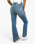 A person wearing blue denim jeans with a straight-to-flare leg, shown from the back against a plain white studio background. The jeans have a light wash and are paired with white sneakers.