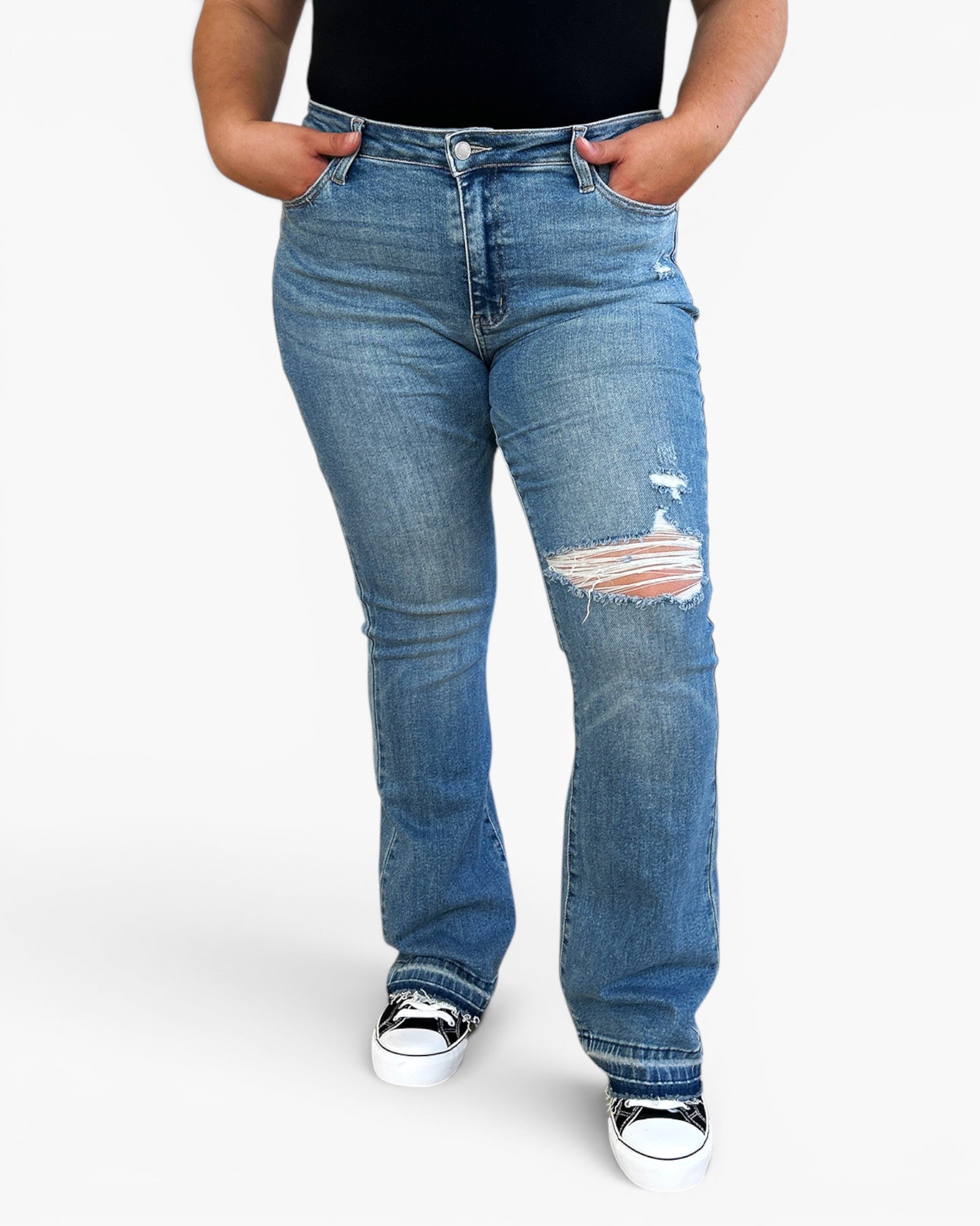 A person wearing medium-wash blue distressed jeans with a ripped knee detail, paired with a black top and black-and-white sneakers, shown against a plain light gray studio background.