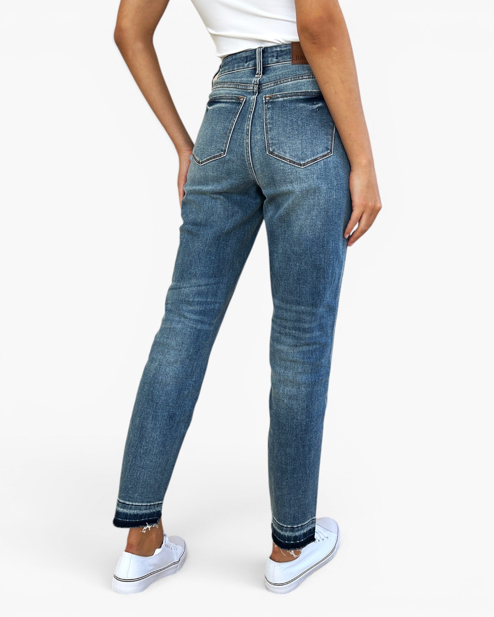 A model shown from the back wearing a pair of high-waisted, ankle-length blue denim jeans with a faded wash and white sneakers, set against a plain white studio background.