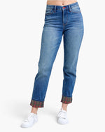A model is wearing a pair of medium wash blue denim jeans with rolled cuffs showing a red and green plaid lining, paired with white sneakers against a plain white background.