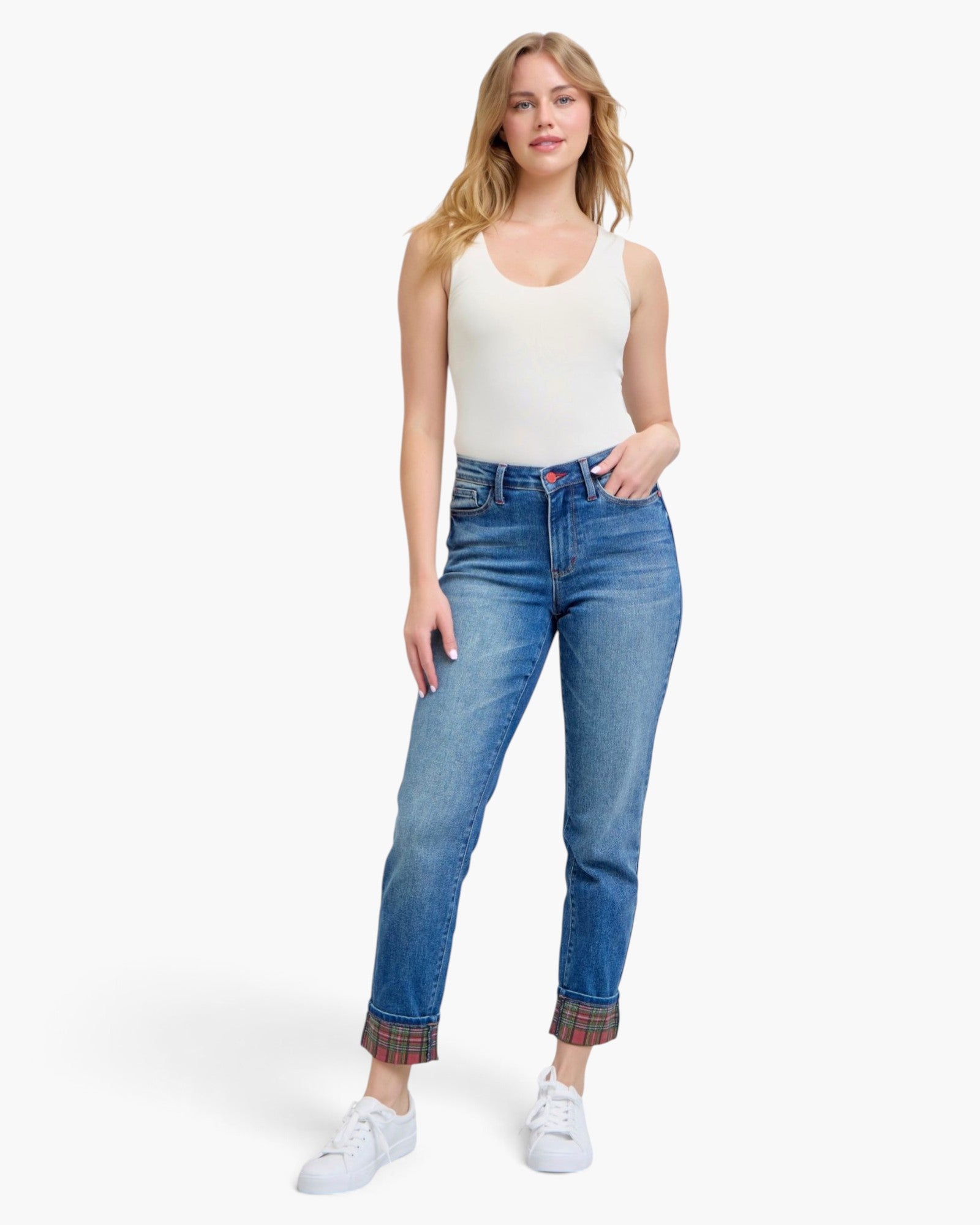 A woman modeling a pair of blue jeans with rolled cuffs showing a plaid lining, paired with a white tank top and white sneakers, against a light gray studio background.
