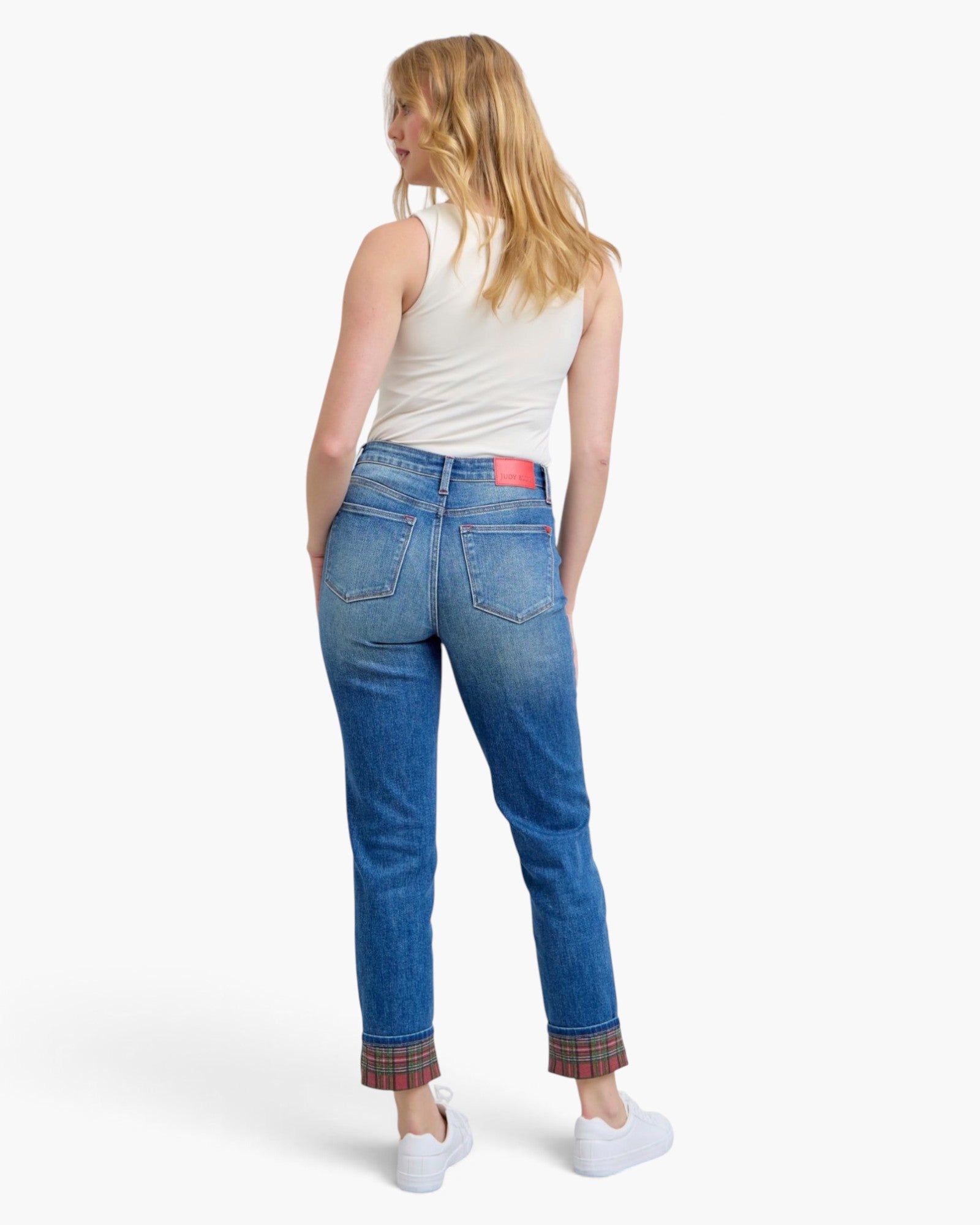 A woman is shown from the back wearing blue jeans with a red plaid cuff lining, a white sleeveless top, and white sneakers against a plain white studio background.