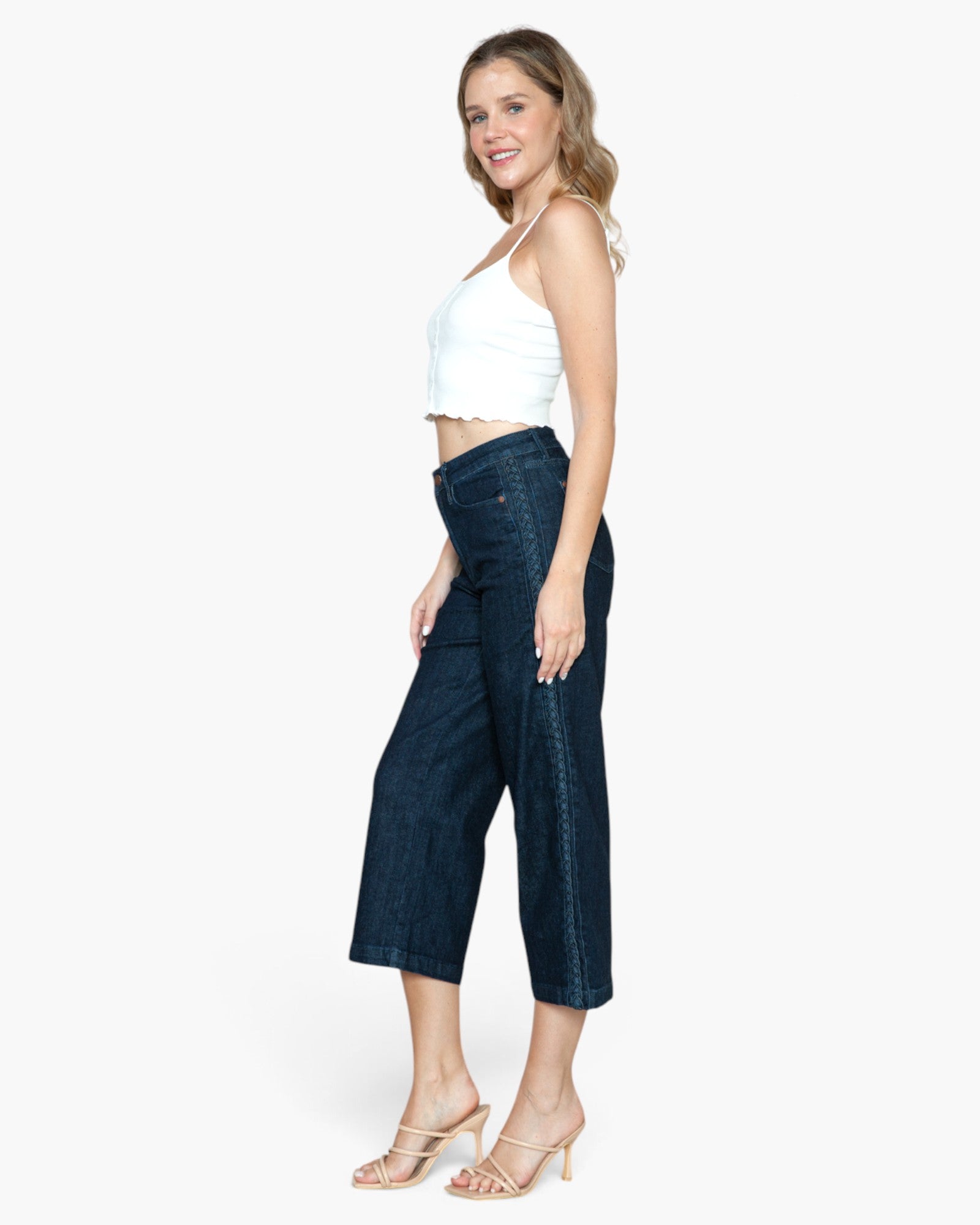 A woman modeling dark blue cropped denim pants with a white halter crop top and beige heeled sandals, shown from the side against a light gray studio background.