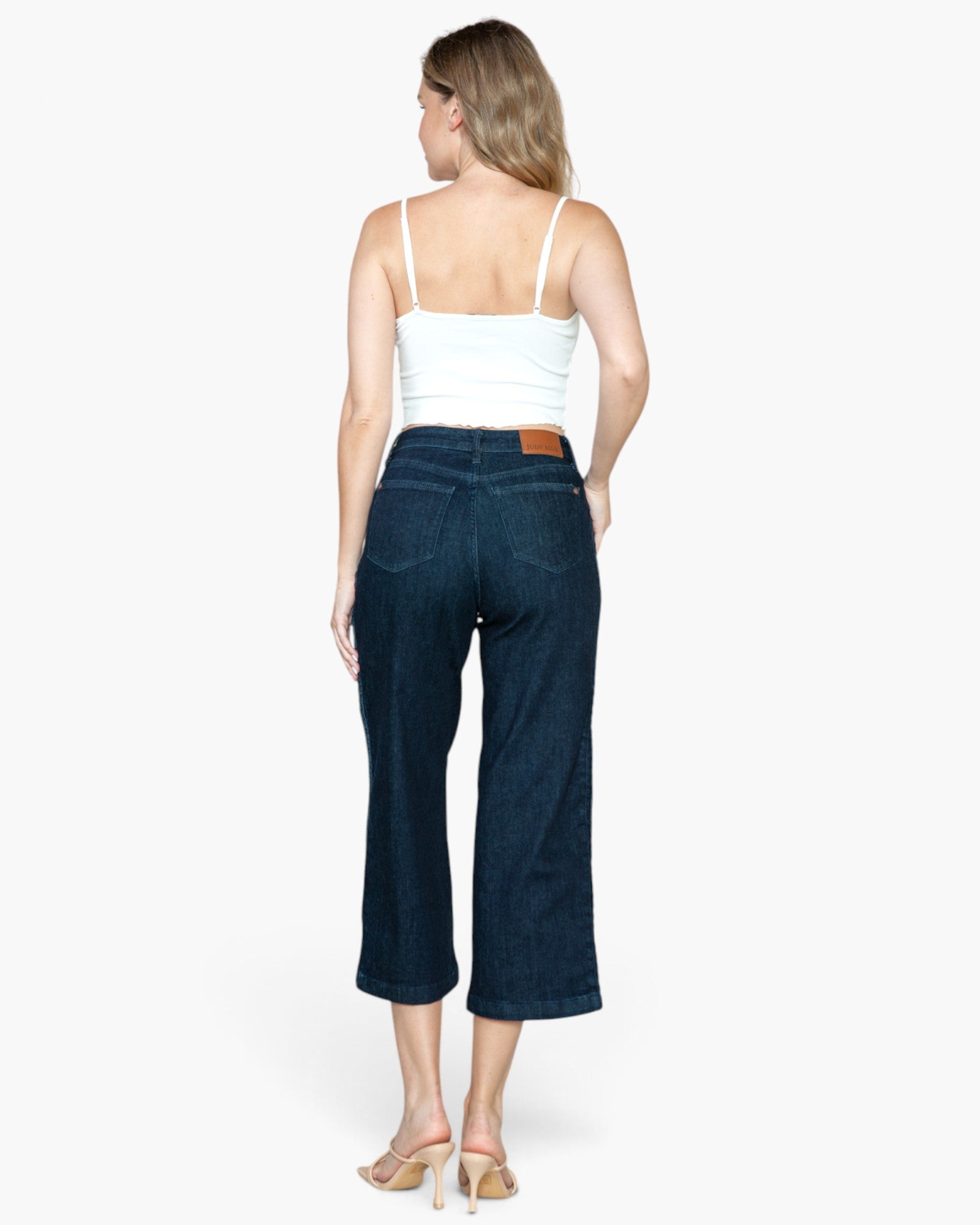 A woman shown from the back wearing dark blue cropped jeans, a white spaghetti-strap top, and beige high-heeled sandals against a light gray studio background.