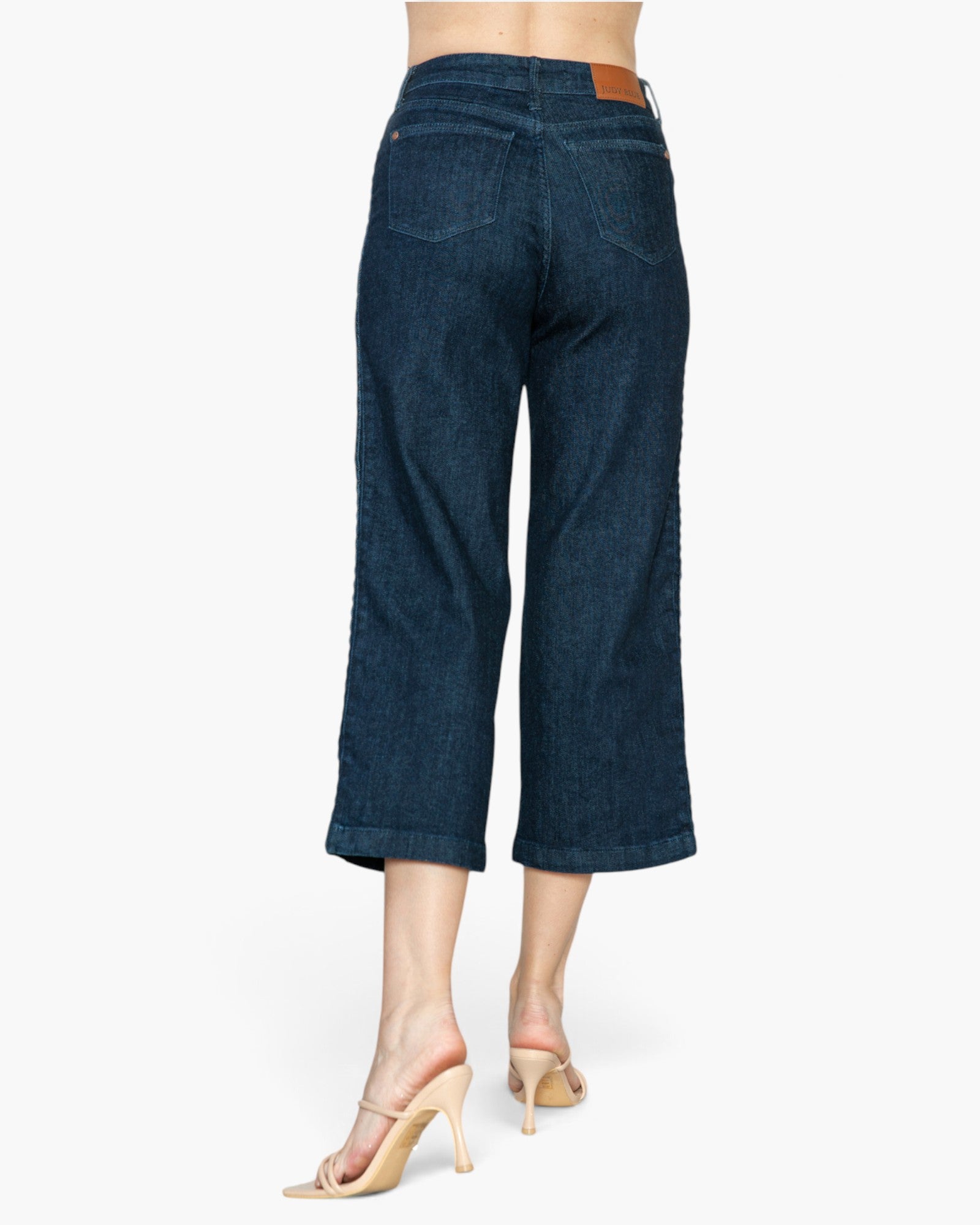 Back view of a model wearing dark blue cropped denim jeans and beige high-heeled sandals against a plain white background.