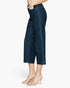 A side view of a model wearing dark blue denim cropped pants with vertical contrast stitching down the outer side seams, paired with beige strappy high-heeled sandals against a plain white background.