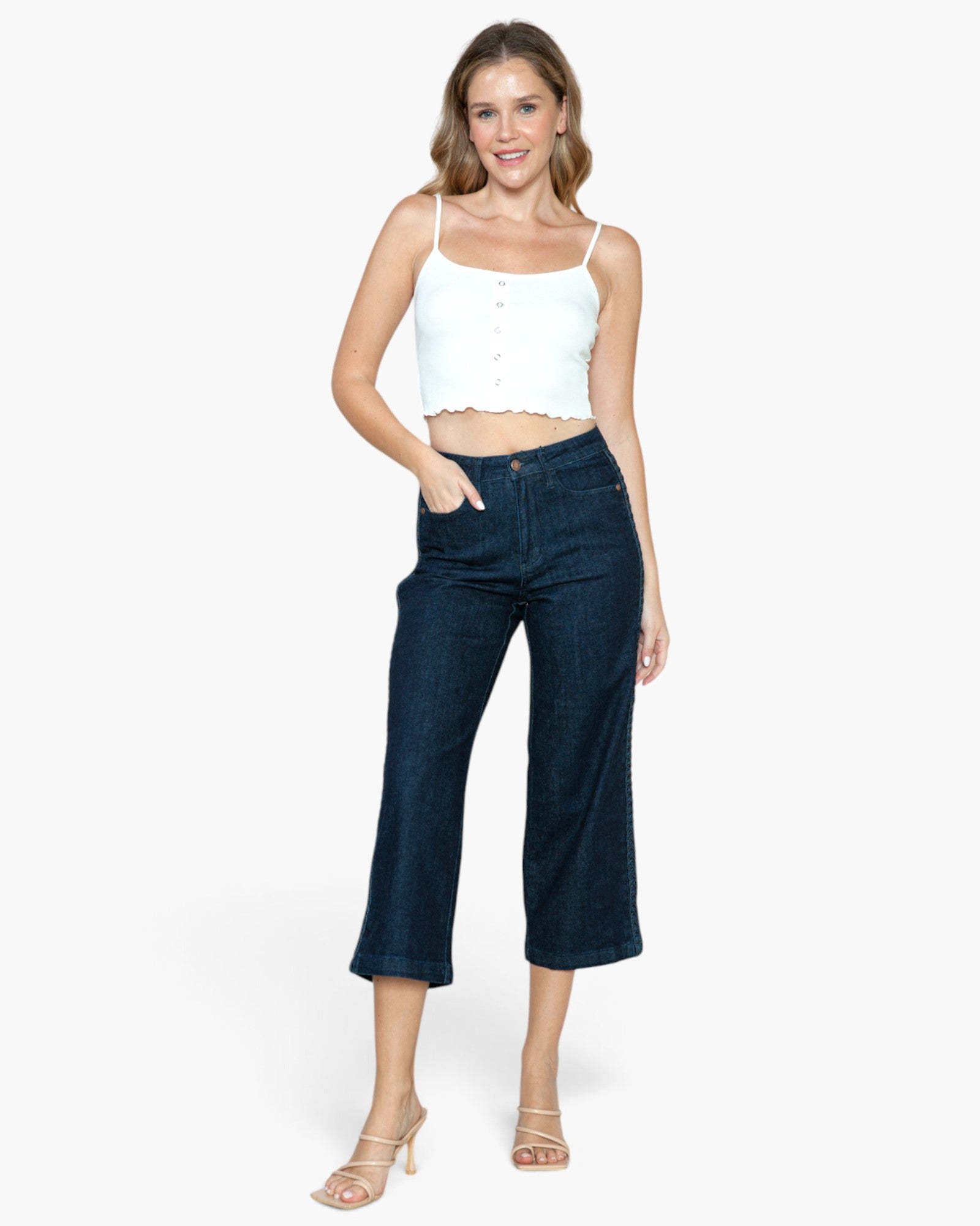 A woman modeling a white cropped camisole top and dark blue cropped denim jeans against a light gray studio background.