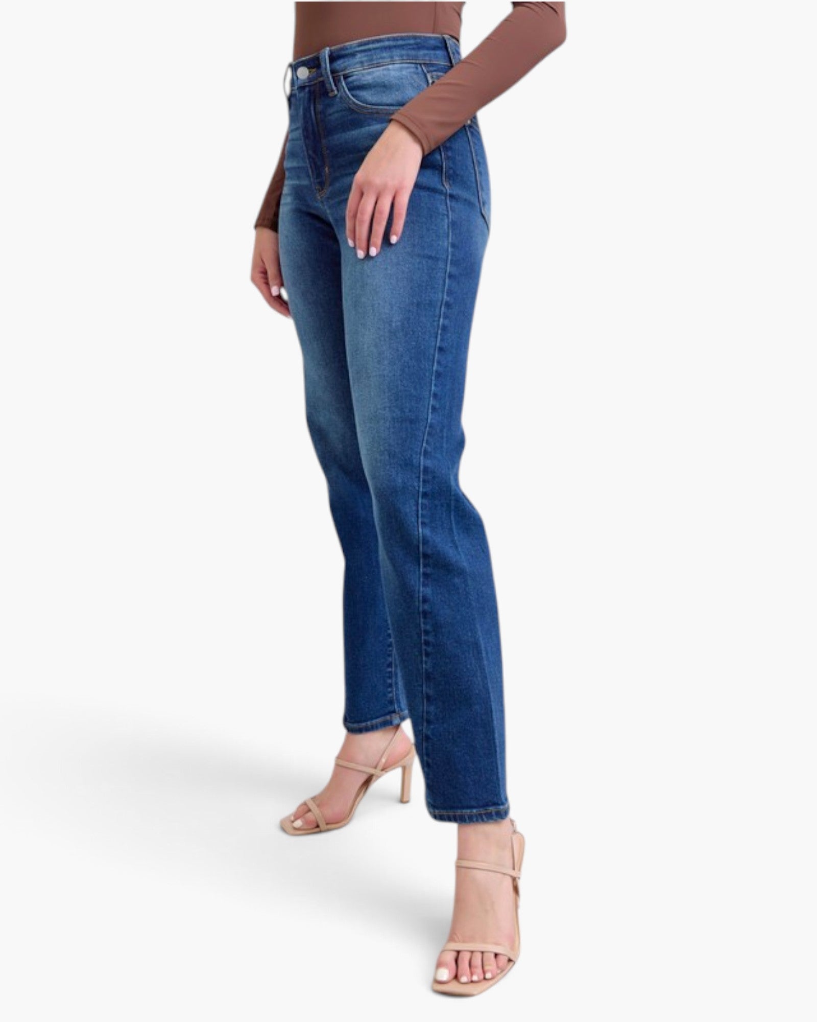 A model wearing a pair of high-waisted, straight-leg blue jeans with a brown long-sleeve top and nude high-heeled sandals, shown from the waist down against a light gray studio background.