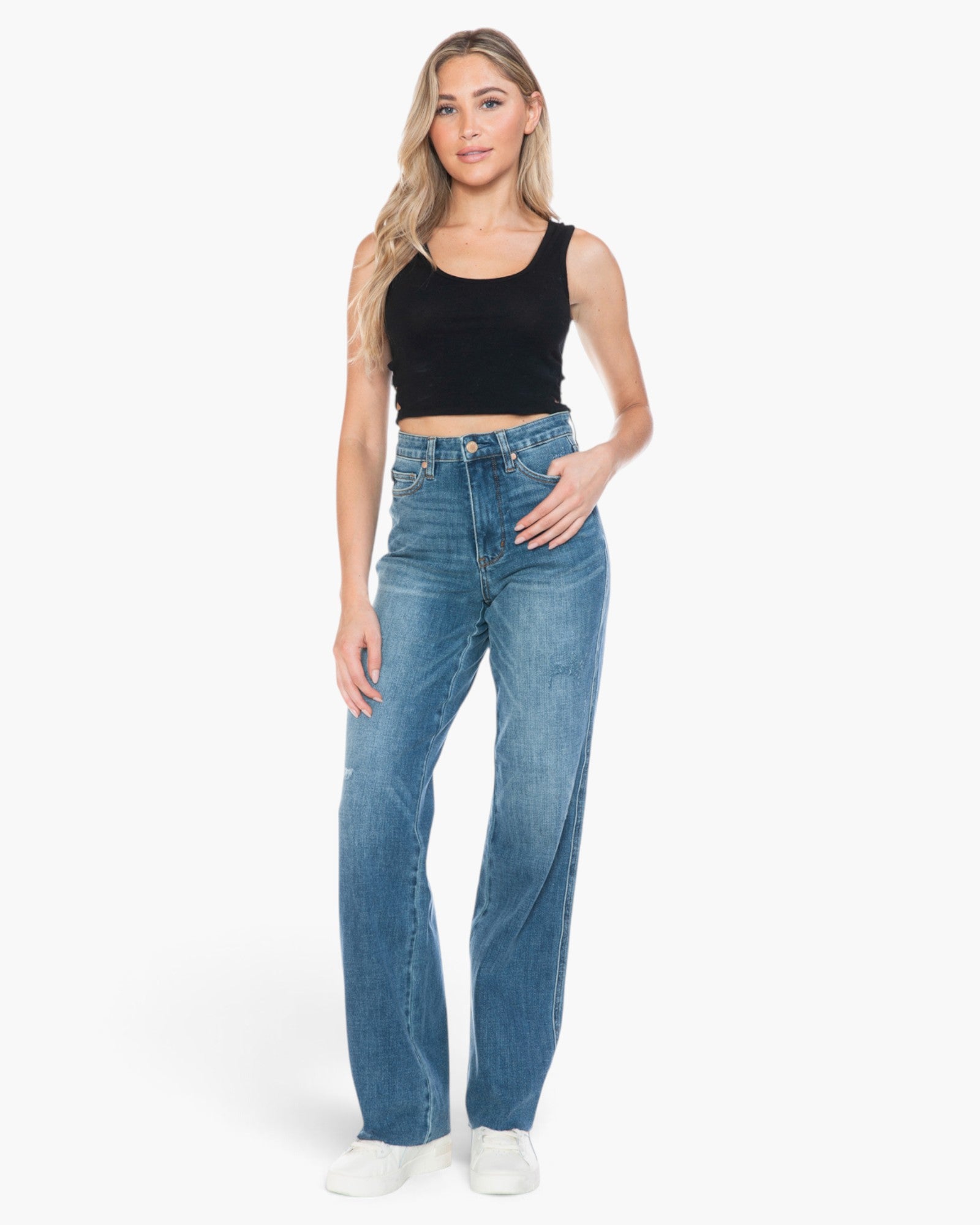 A woman modeling a pair of high-waisted, wide-leg blue jeans with a black crop tank top and white sneakers, standing against a plain white studio background.