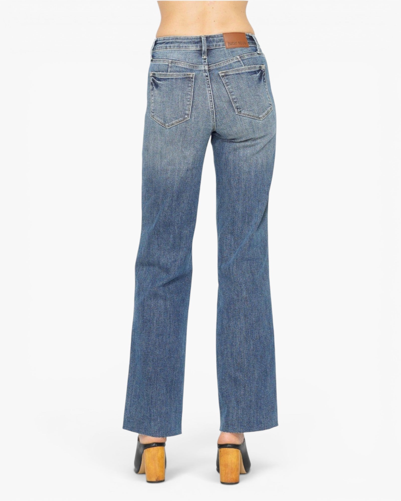 Rear view of a person wearing blue denim jeans with a straight-to-medium leg fit, paired with black and tan block-heel sandals, against a plain white studio background.