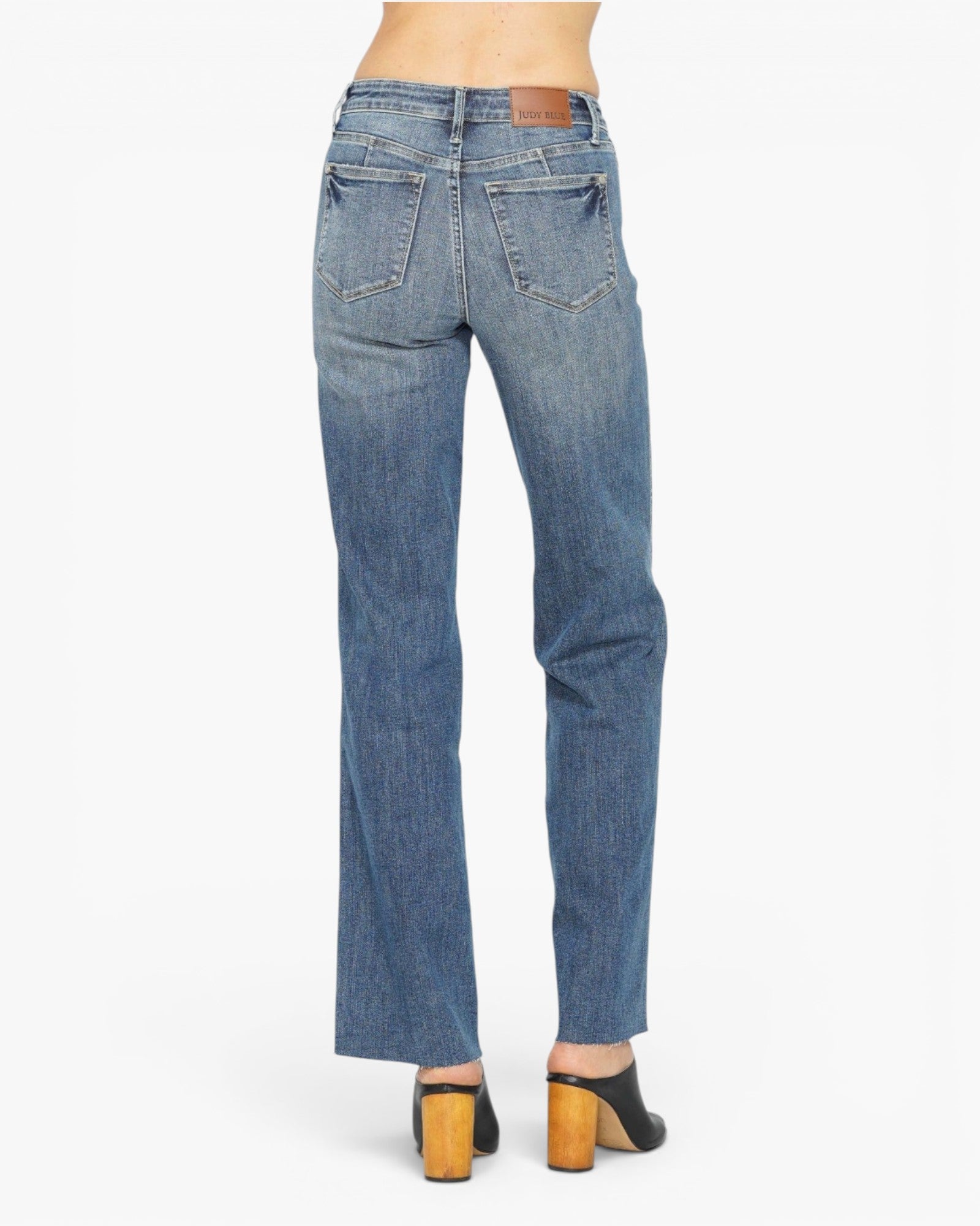 Rear view of a model wearing medium-wash blue denim jeans with a straight-to-slightly-flared leg, paired with black mule heels with wooden block heels, against a plain white background.