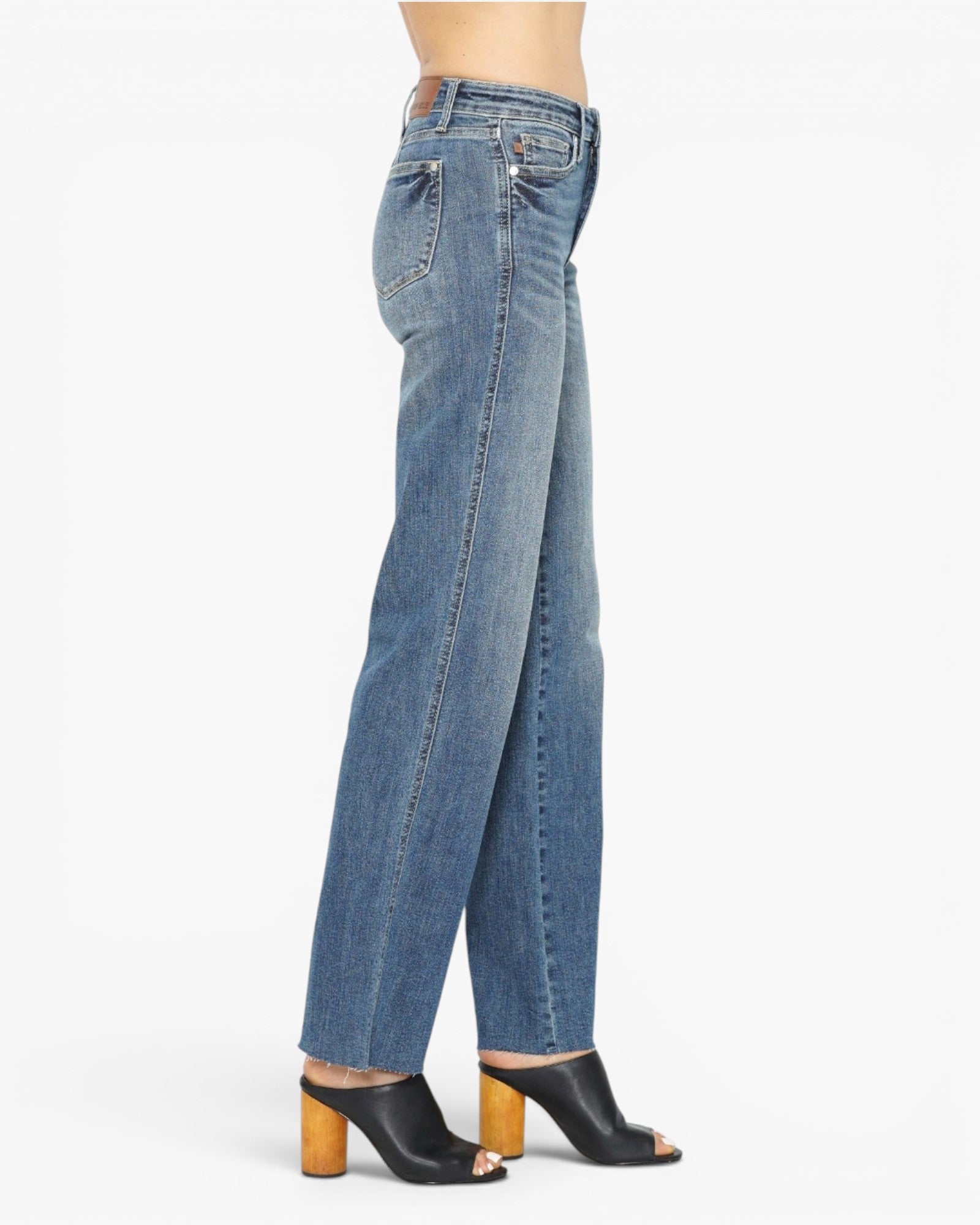 A side profile view of a model wearing medium-wash blue denim jeans and black peep-toe mule heels with wooden block heels, set against a plain white studio background.