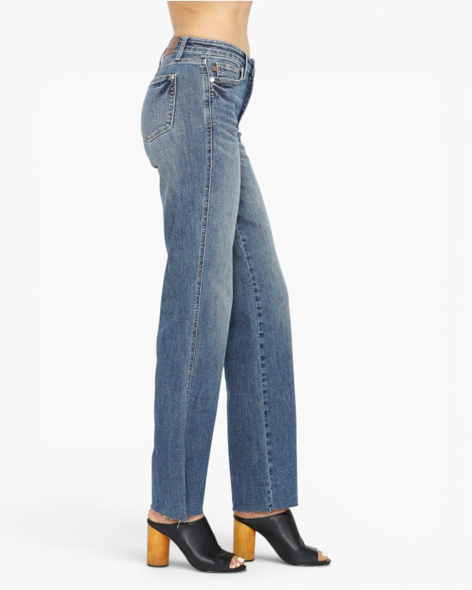 A side profile view of a model wearing medium-wash blue denim jeans and black peep-toe mule heels with wooden block heels, set against a plain white studio background.