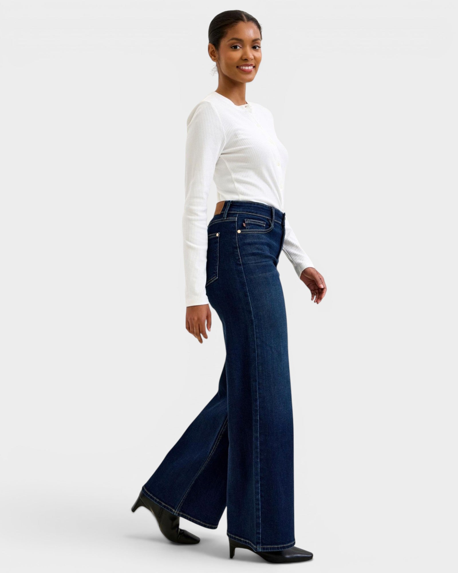 Woman wearing a white long-sleeve top and dark blue flared jeans on a light gray background