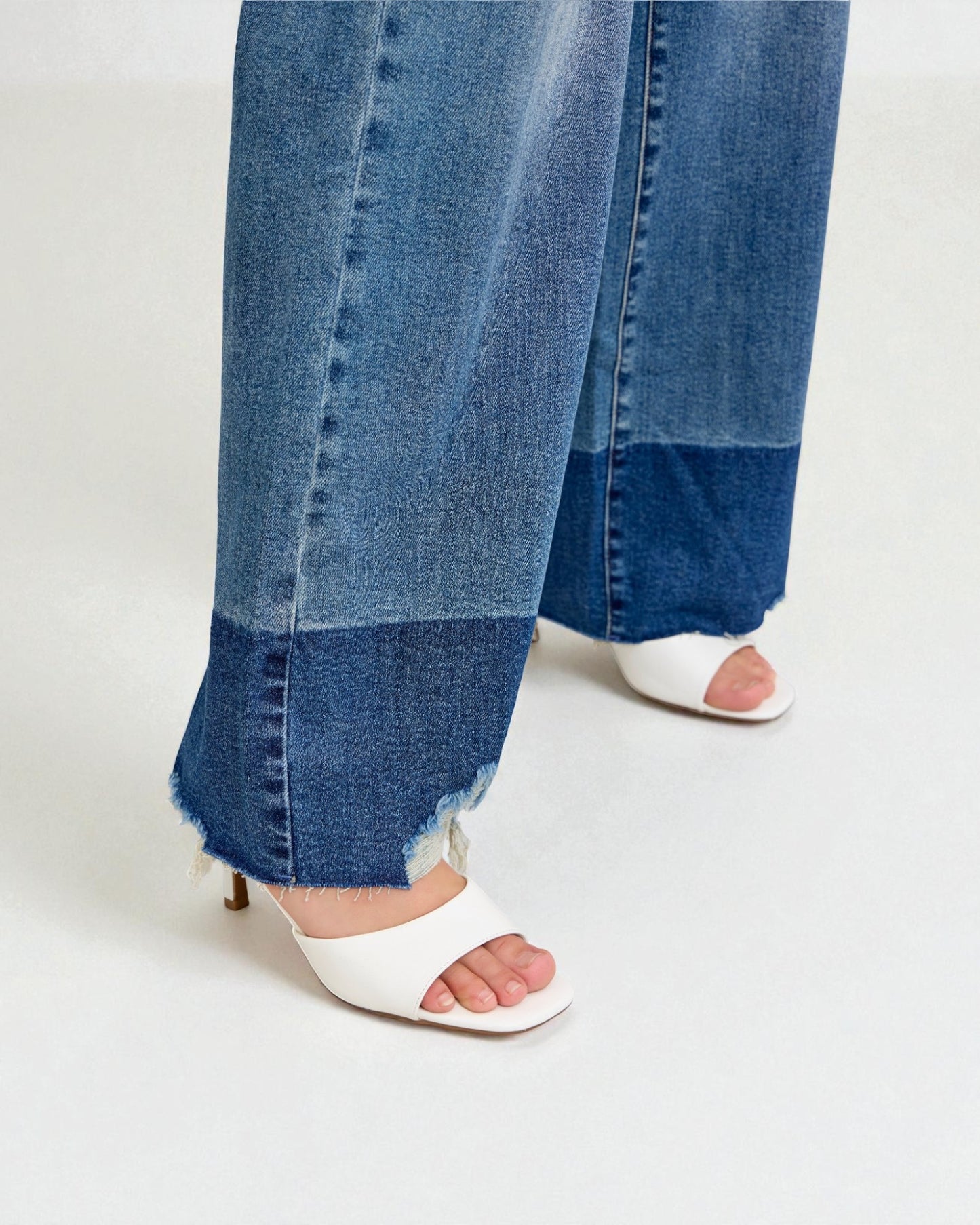 Close-up of blue jeans with a frayed hem and white open-toe shoes on a light gray background