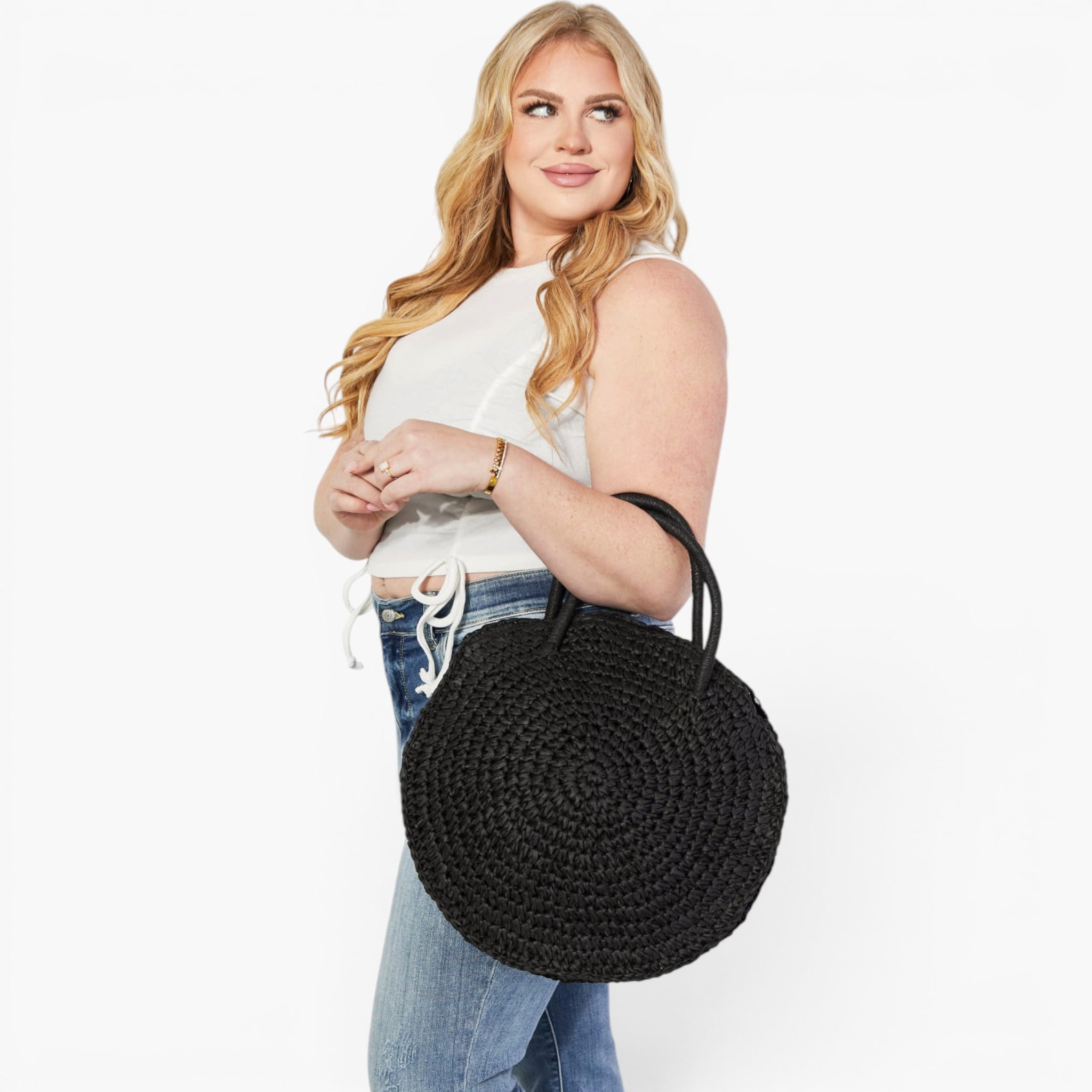 A woman wearing a white sleeveless crop top and blue jeans carries a large round black woven straw bag with two top handles against a plain white background.