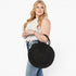A woman wearing a white sleeveless crop top and blue jeans carries a large round black woven straw bag with two top handles against a plain white background.