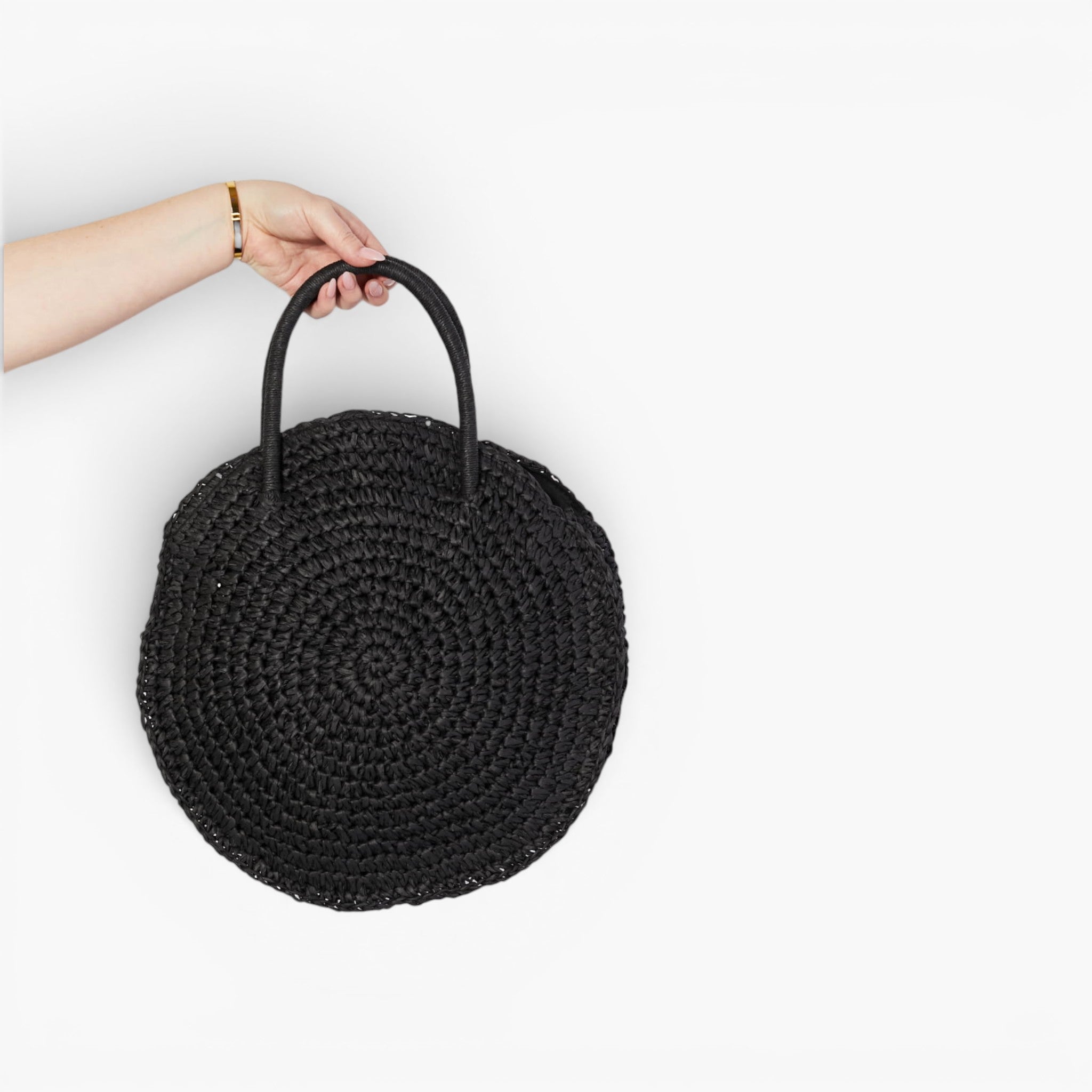 A hand holding a round black woven straw bag with two top handles against a plain white background.