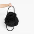 A black textured woven handbag with black handles, held by a person's hand against a plain white background.