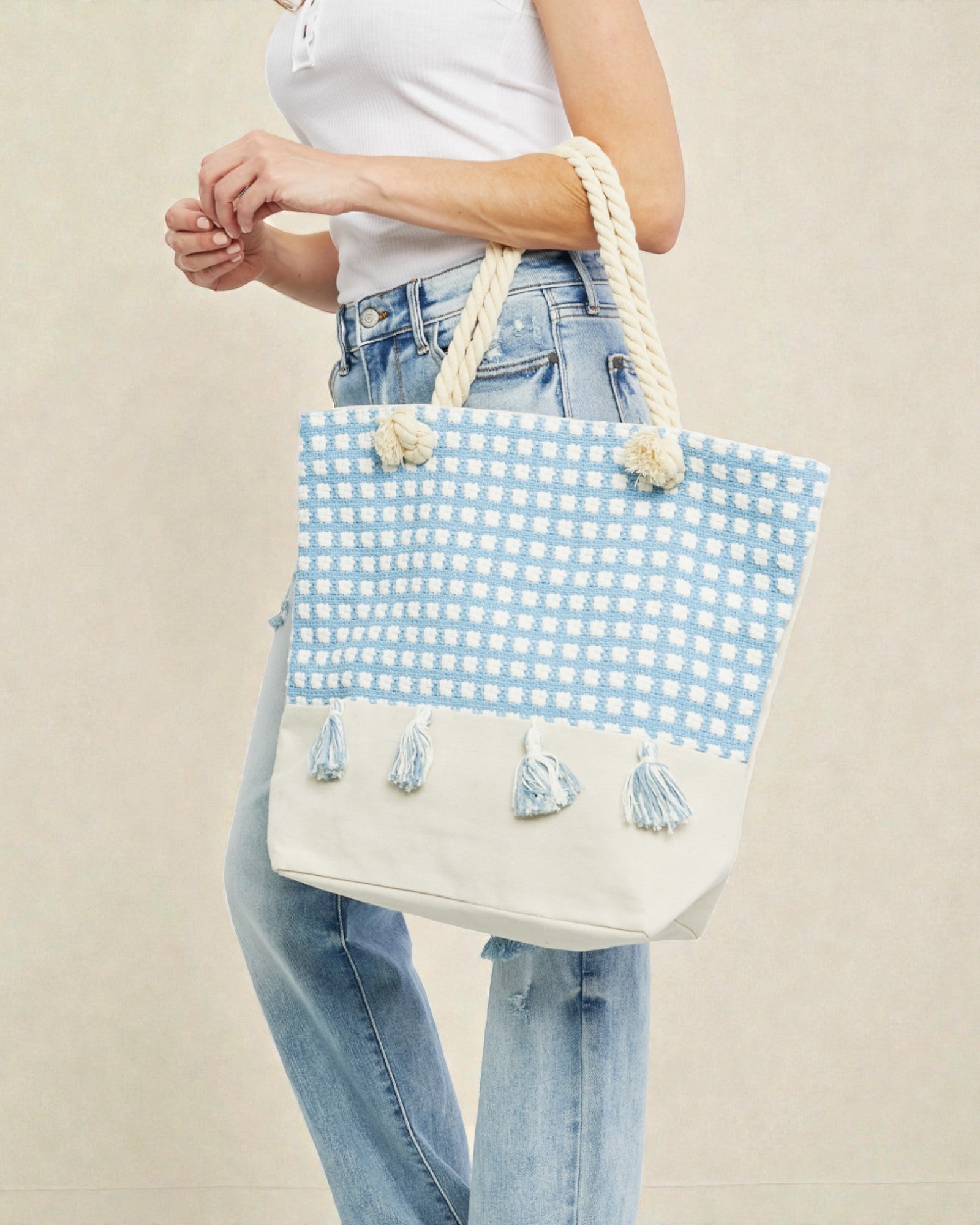 Person holding a blue and white checkered tote bag with tassels on a beige background
