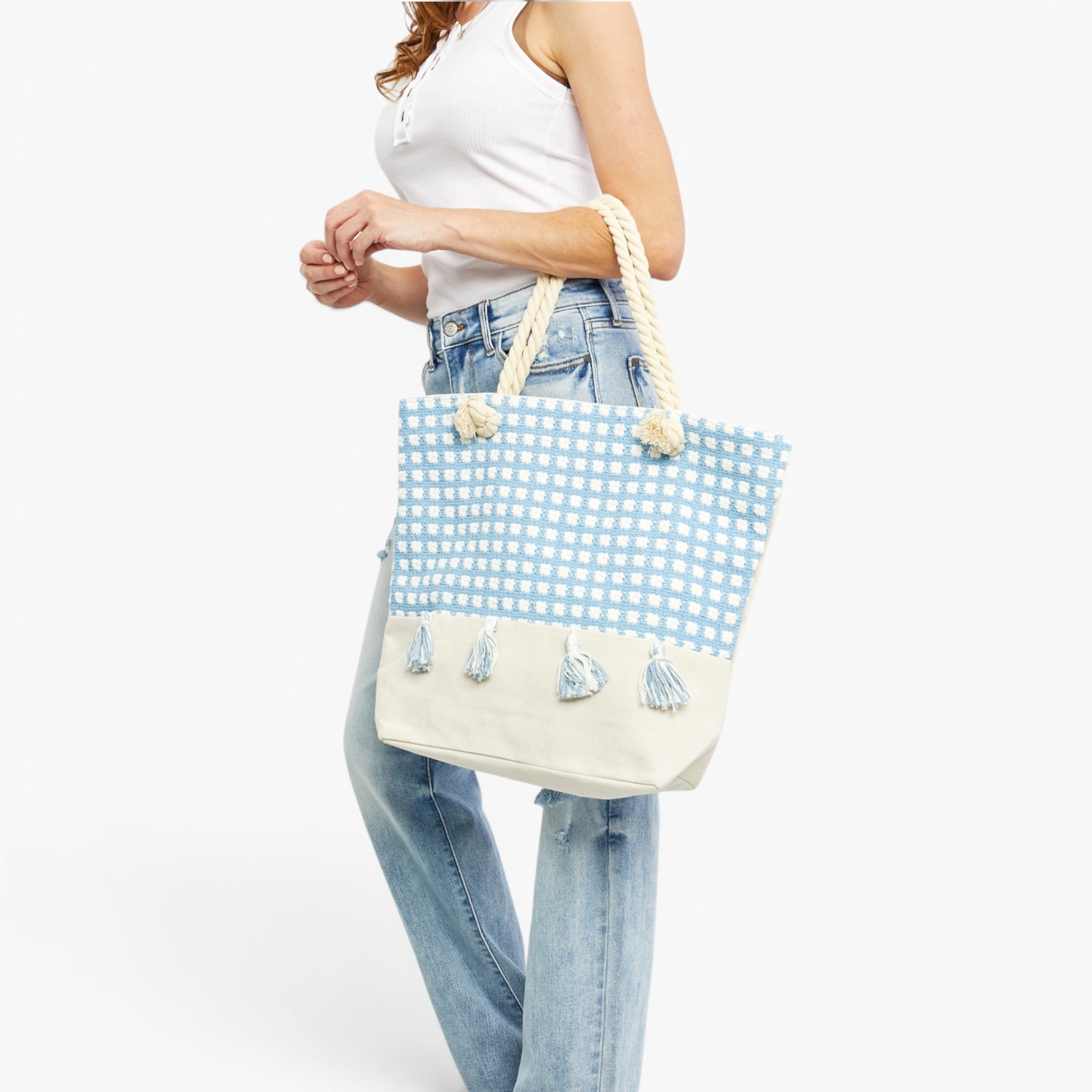 A woman wearing a white tank top and light blue jeans carries a large tote bag with a light blue and white gingham pattern on the upper half, a solid cream lower half with tassels, and thick cream rope handles.