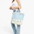 A woman wearing a white tank top and light blue jeans carries a large tote bag with a light blue and white gingham pattern on the upper half, a solid cream lower half with tassels, and thick cream rope handles.