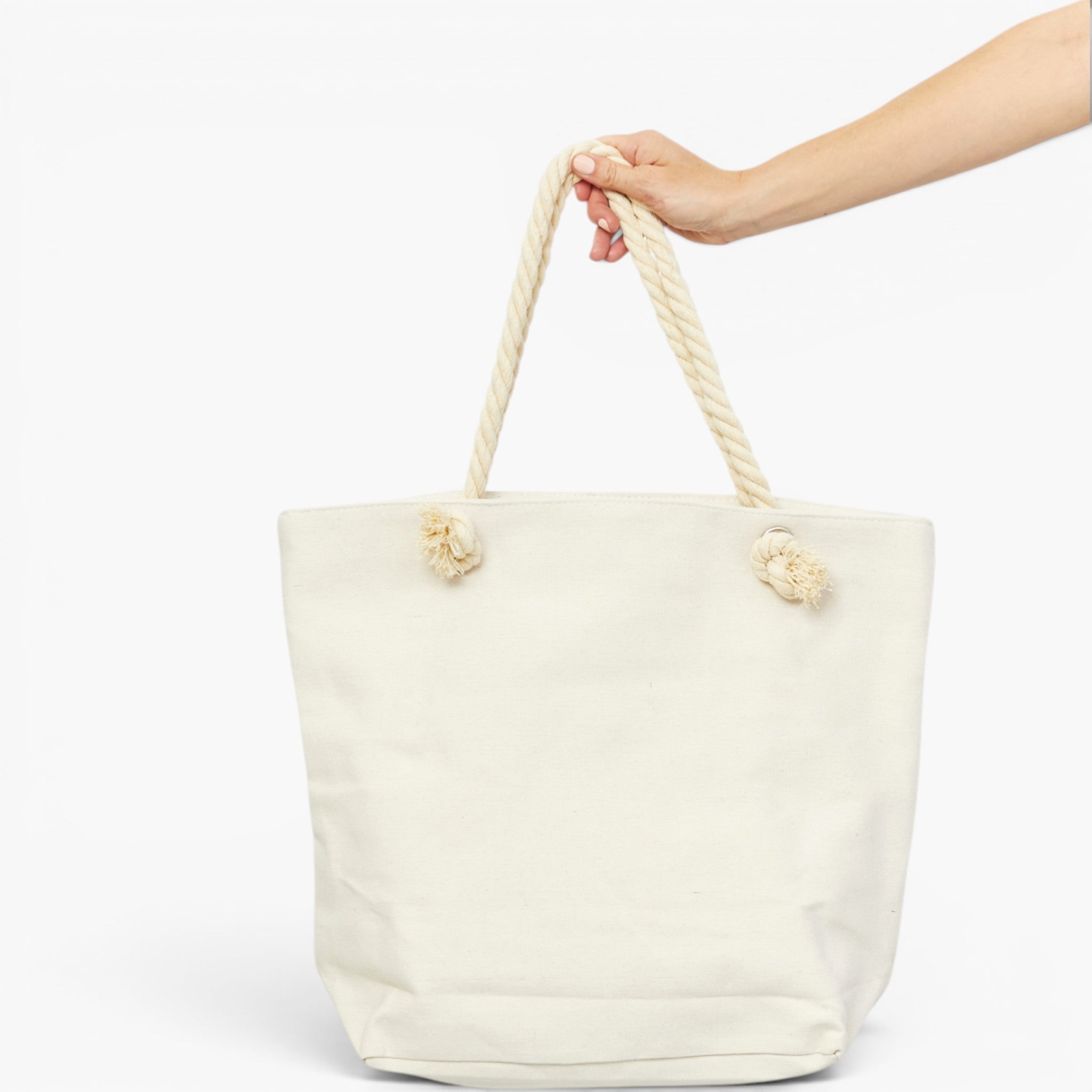 An off-white canvas tote bag with thick rope handles knotted at the sides, held by a person's hand against a plain white background.