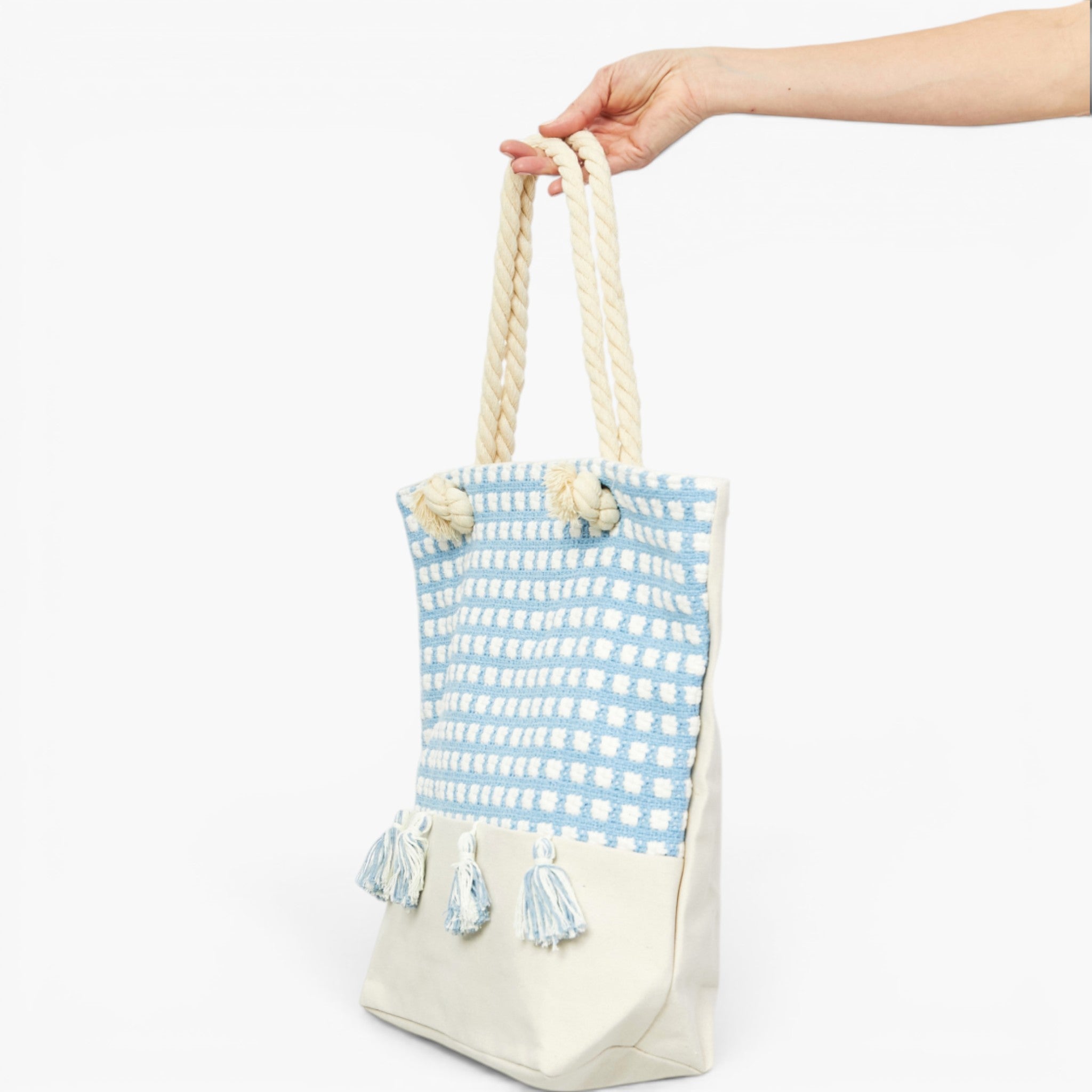 A woven tote bag with a light blue and white patterned upper half, cream-colored bottom half, and thick cream rope handles, held by a hand against a plain white background.