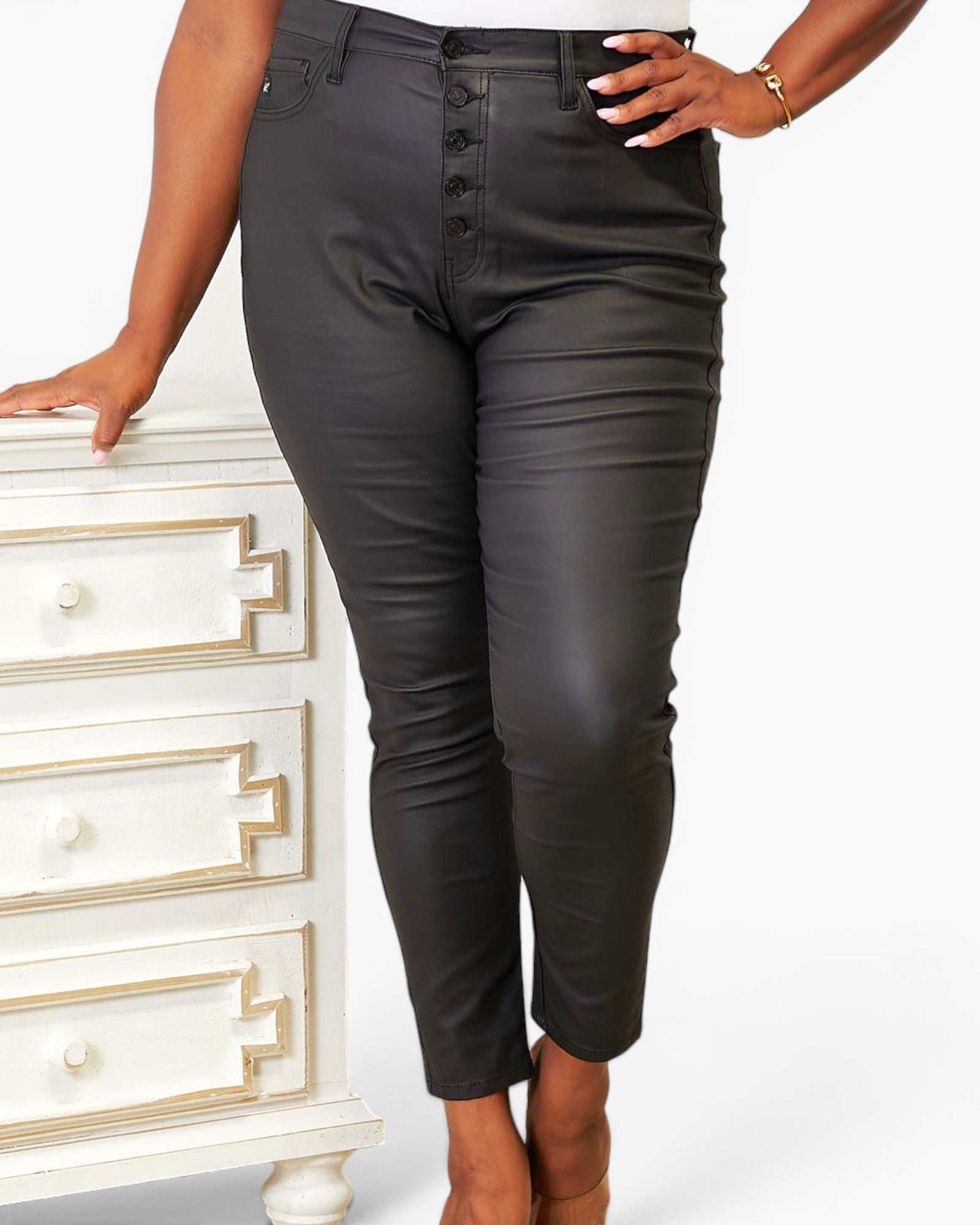 A model wearing black faux-leather cropped pants with a button-fly front, standing next to a white dresser.