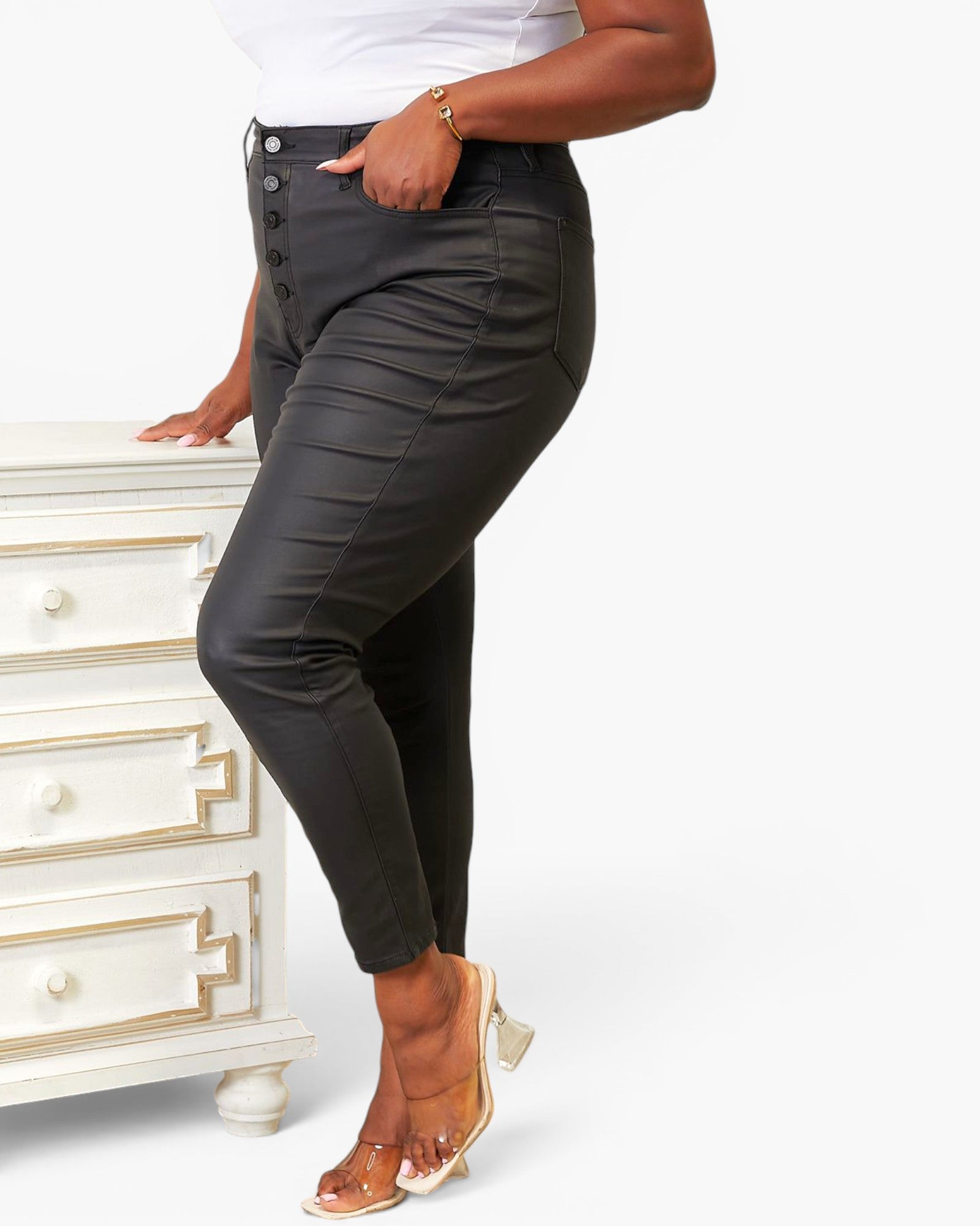 A model wearing black high-waisted faux-leather pants with a button fly, standing next to a white dresser against a white studio background.