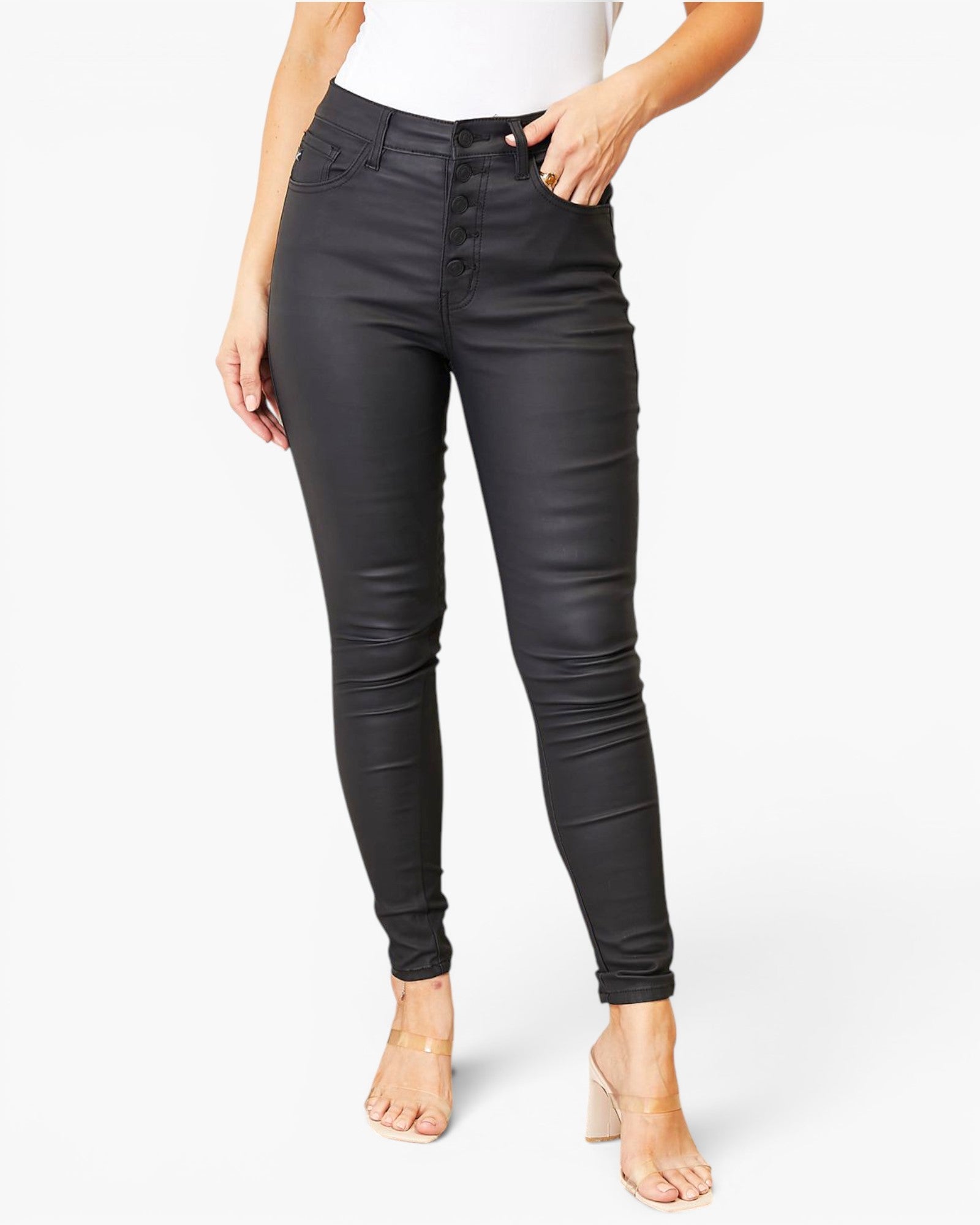 A model wearing a pair of black high-waisted, slim-fit skinny jeans with a glossy finish, paired with a white top and beige high-heeled sandals, against a plain white studio background.