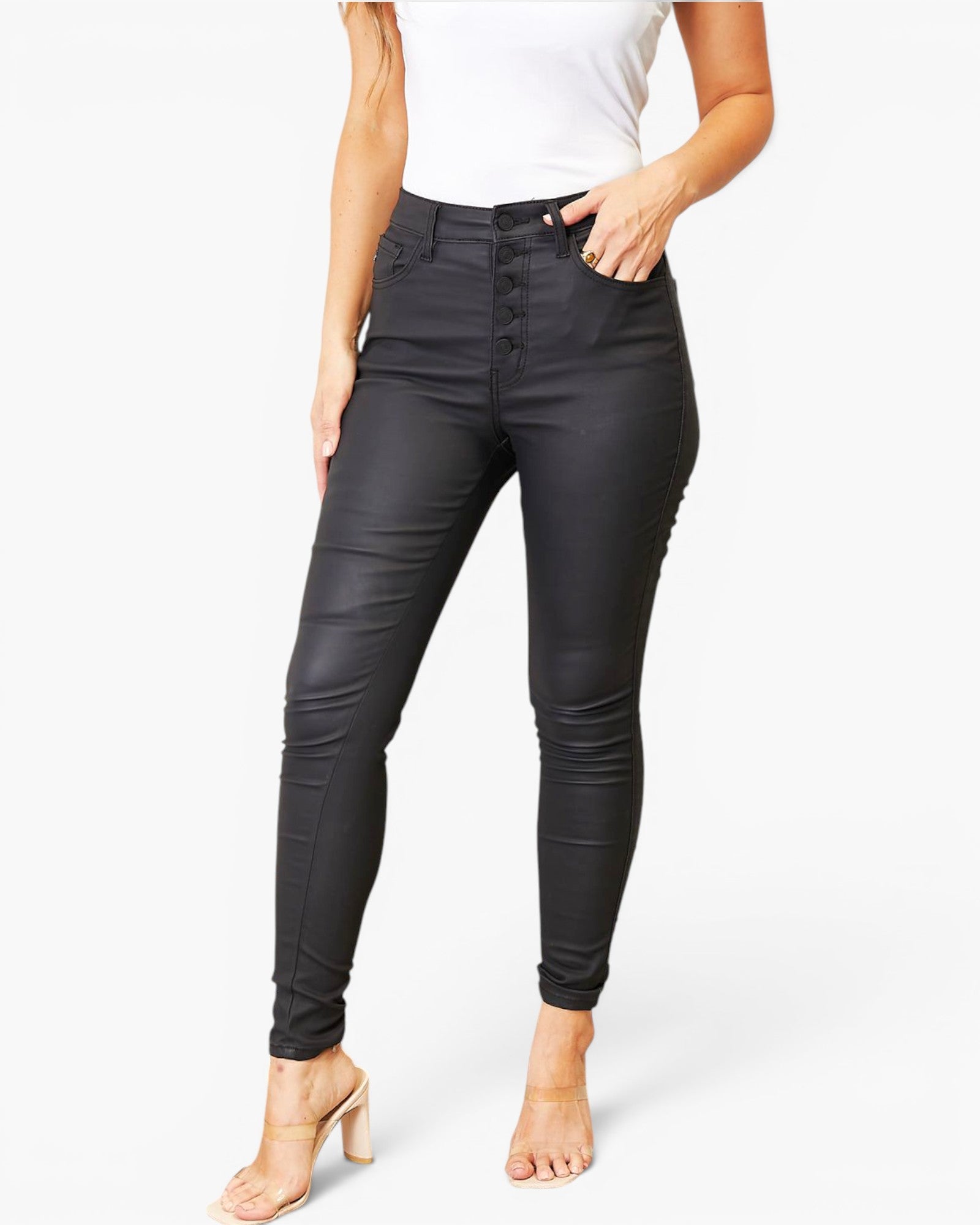 A model wearing black high-waisted faux-leather skinny jeans with a button-fly front, styled with a white top and beige high-heeled sandals against a light gray background.