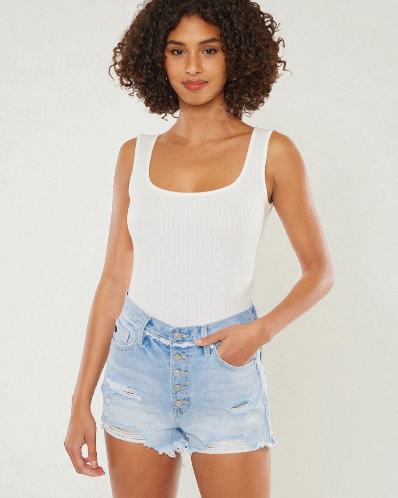 Woman wearing a white tank top and light blue denim shorts on a plain background