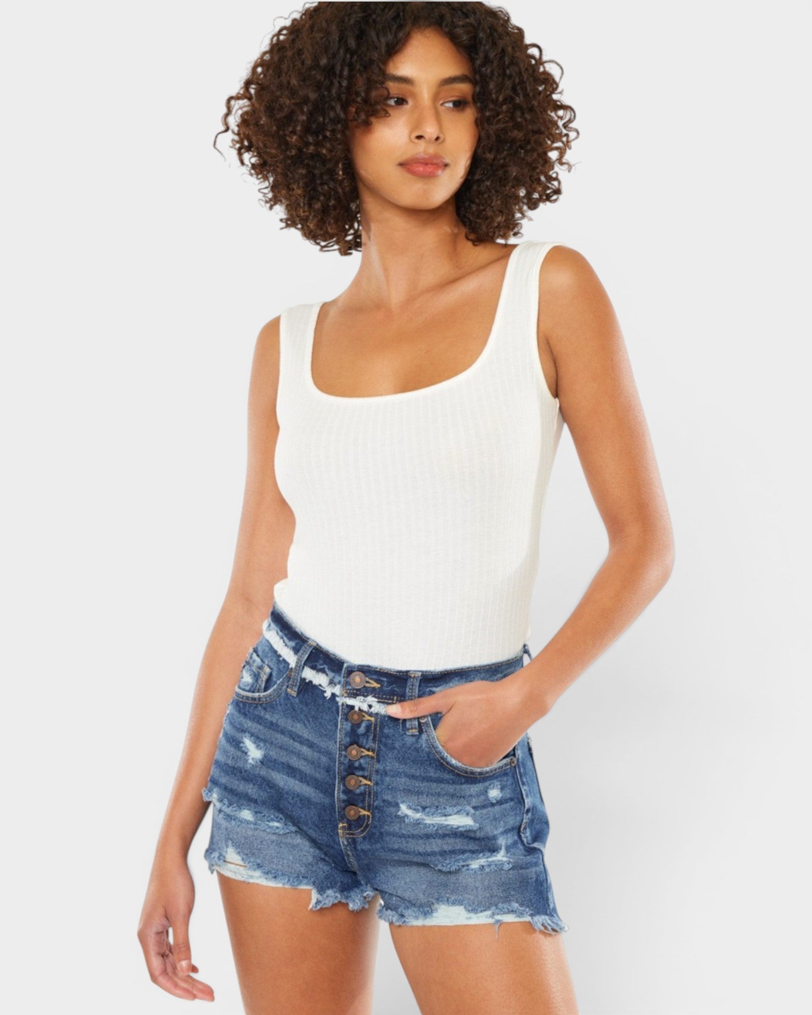 A woman modeling a white ribbed tank top and distressed blue denim shorts with a button fly, set against a plain light gray studio background.