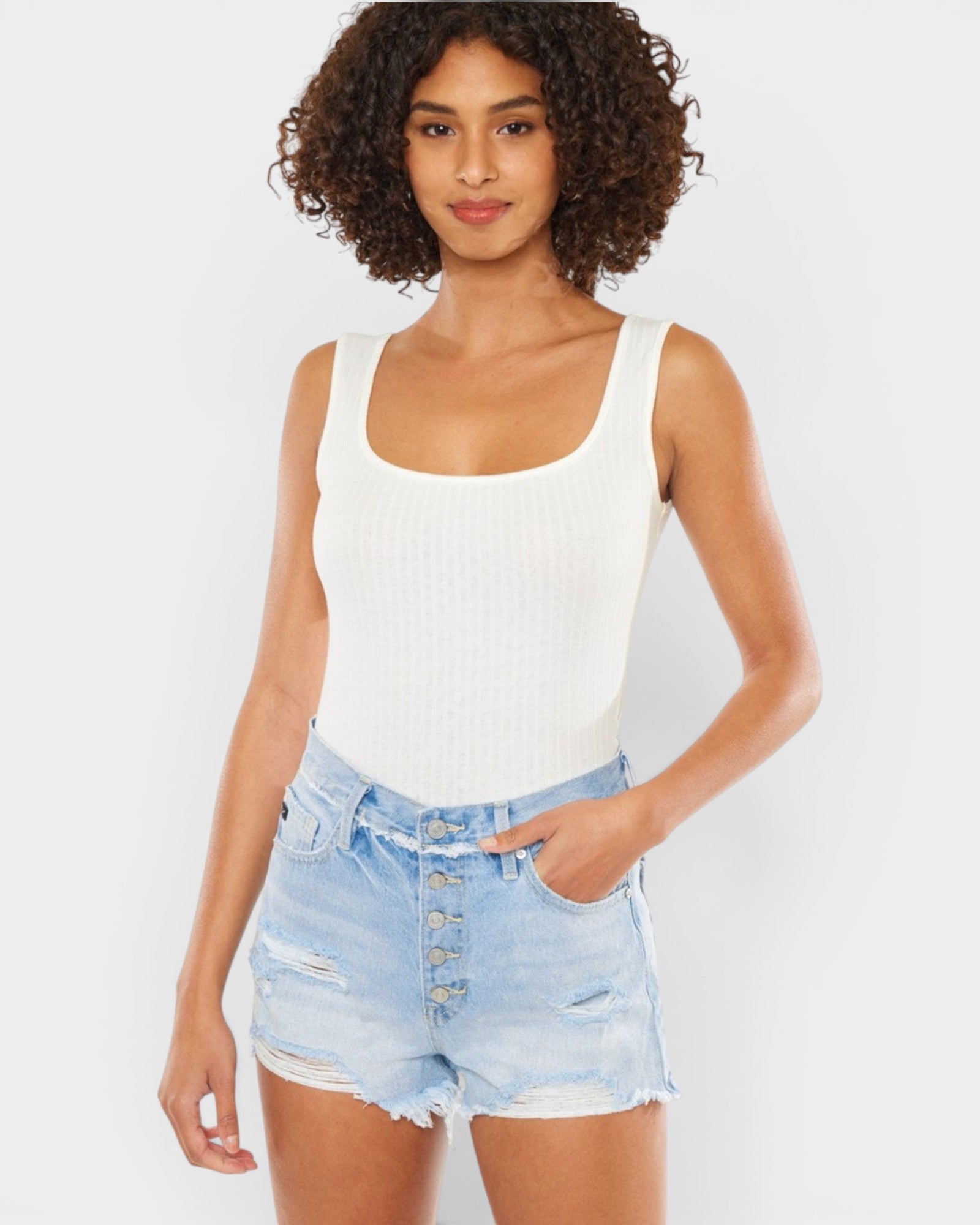 A woman modeling a white ribbed tank top and light blue distressed denim shorts against a plain light gray studio background.