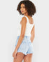 A woman with curly brown hair is shown from the back wearing a white ribbed tank top and light blue denim shorts with a frayed hem, set against a plain light gray background.