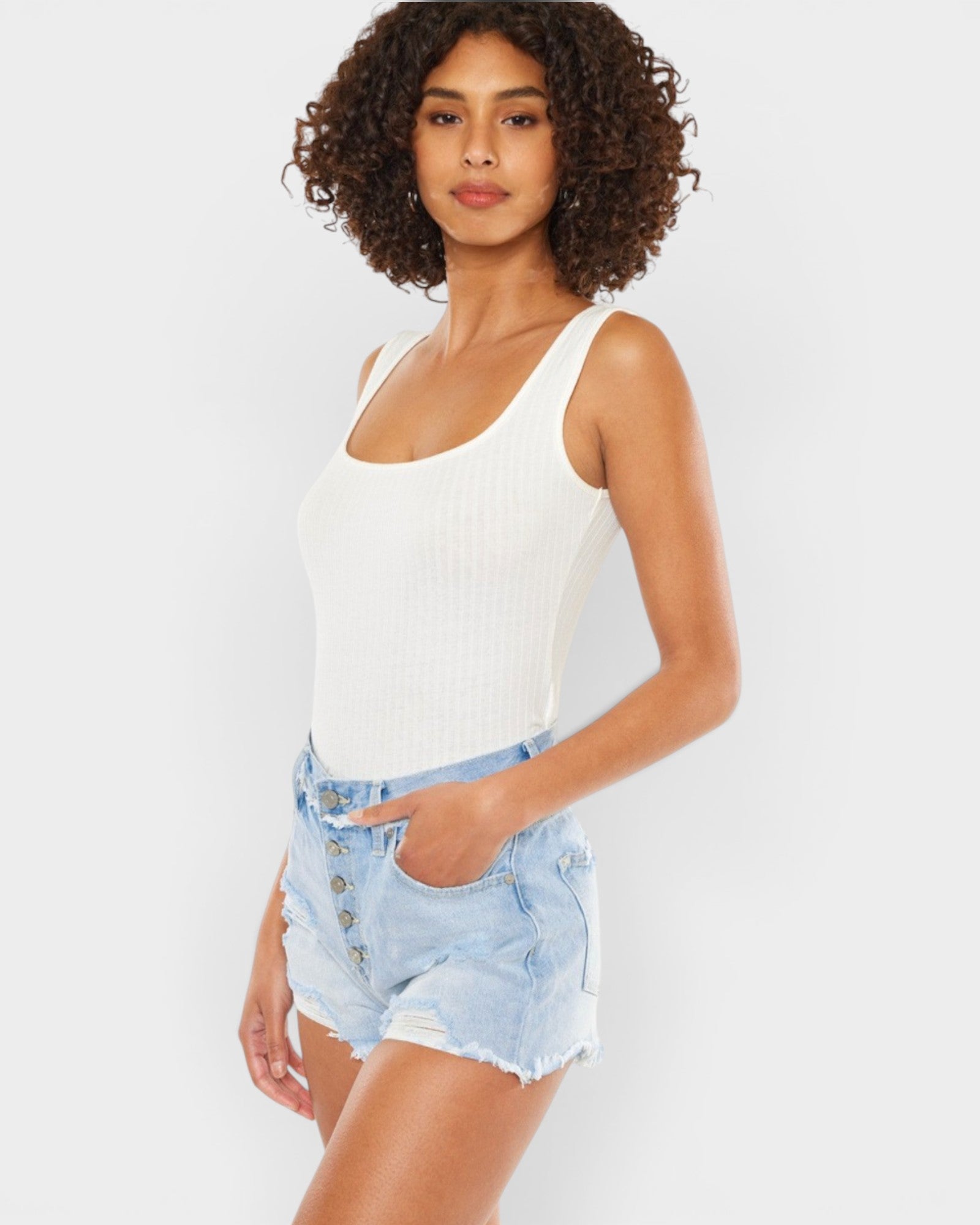 A woman modeling a white ribbed scoop-neck tank top paired with light-wash denim shorts against a light gray studio background.