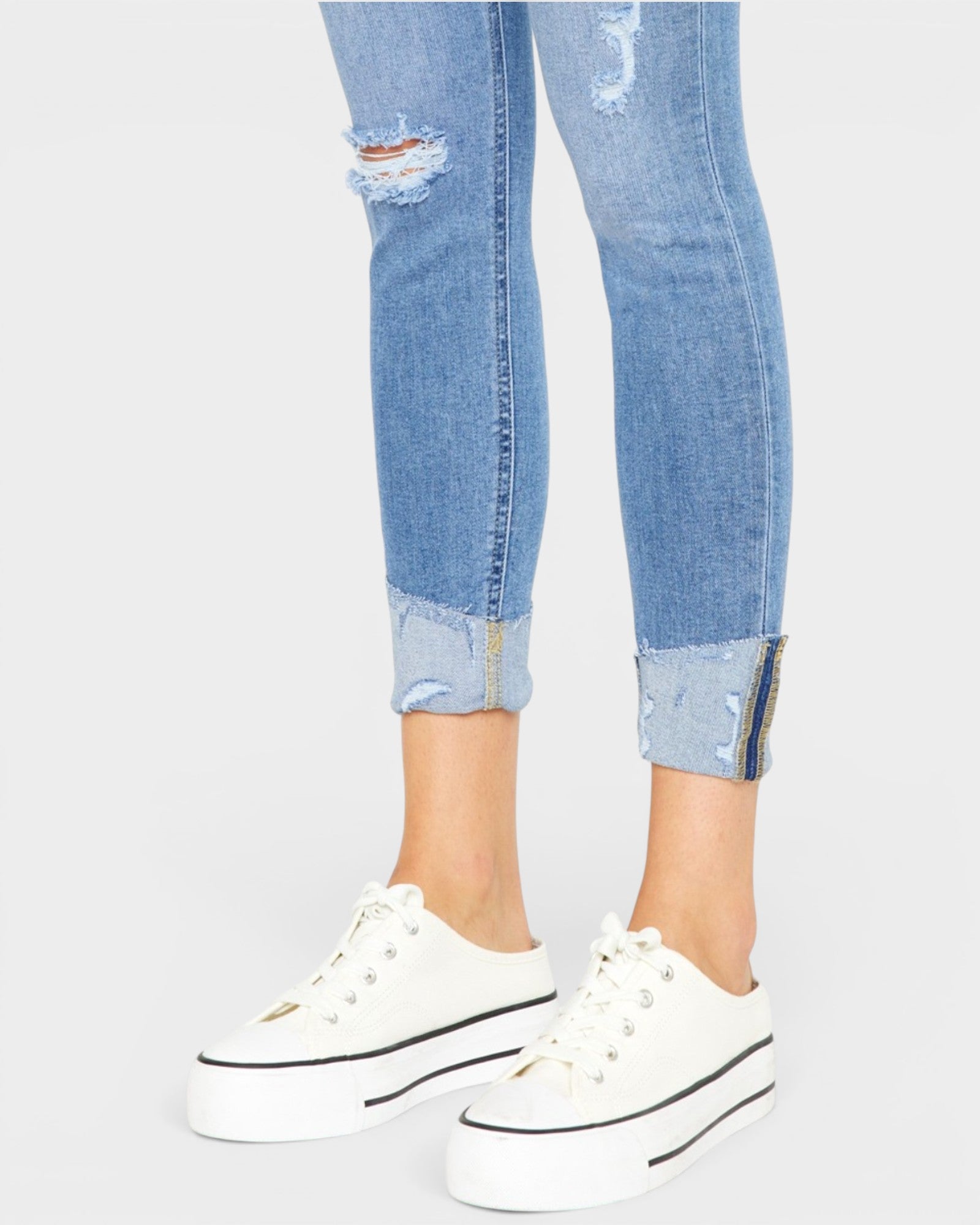 Ripped blue jeans with rolled-up cuffs worn with white sneakers on a light gray background
