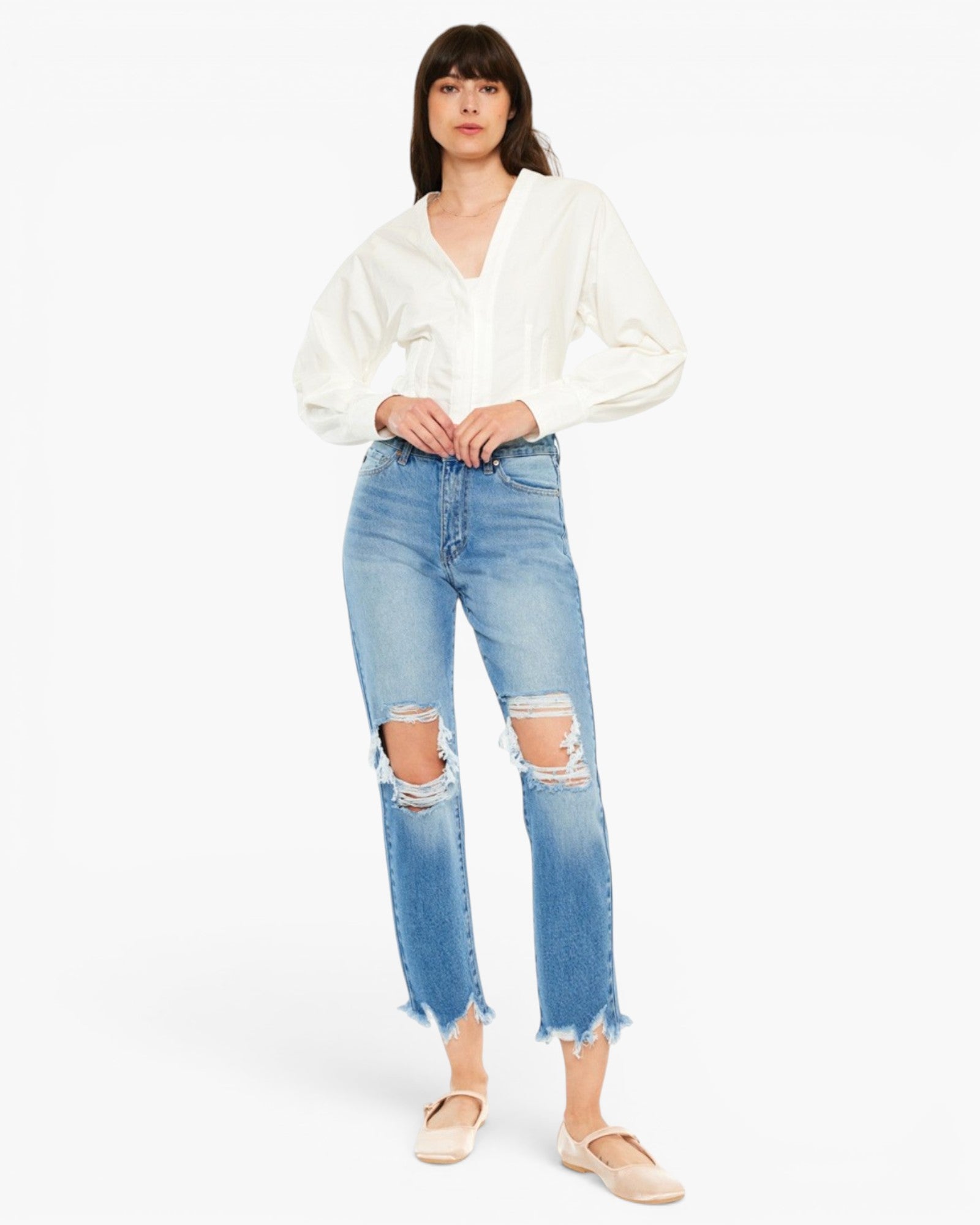 A model wearing a white long-sleeve blouse and light-wash blue distressed cropped jeans with frayed hems, paired with beige flats, against a plain white studio background.