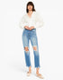 A model wearing a white long-sleeve blouse and light-wash blue distressed cropped jeans with frayed hems, paired with beige flats, against a plain white studio background.