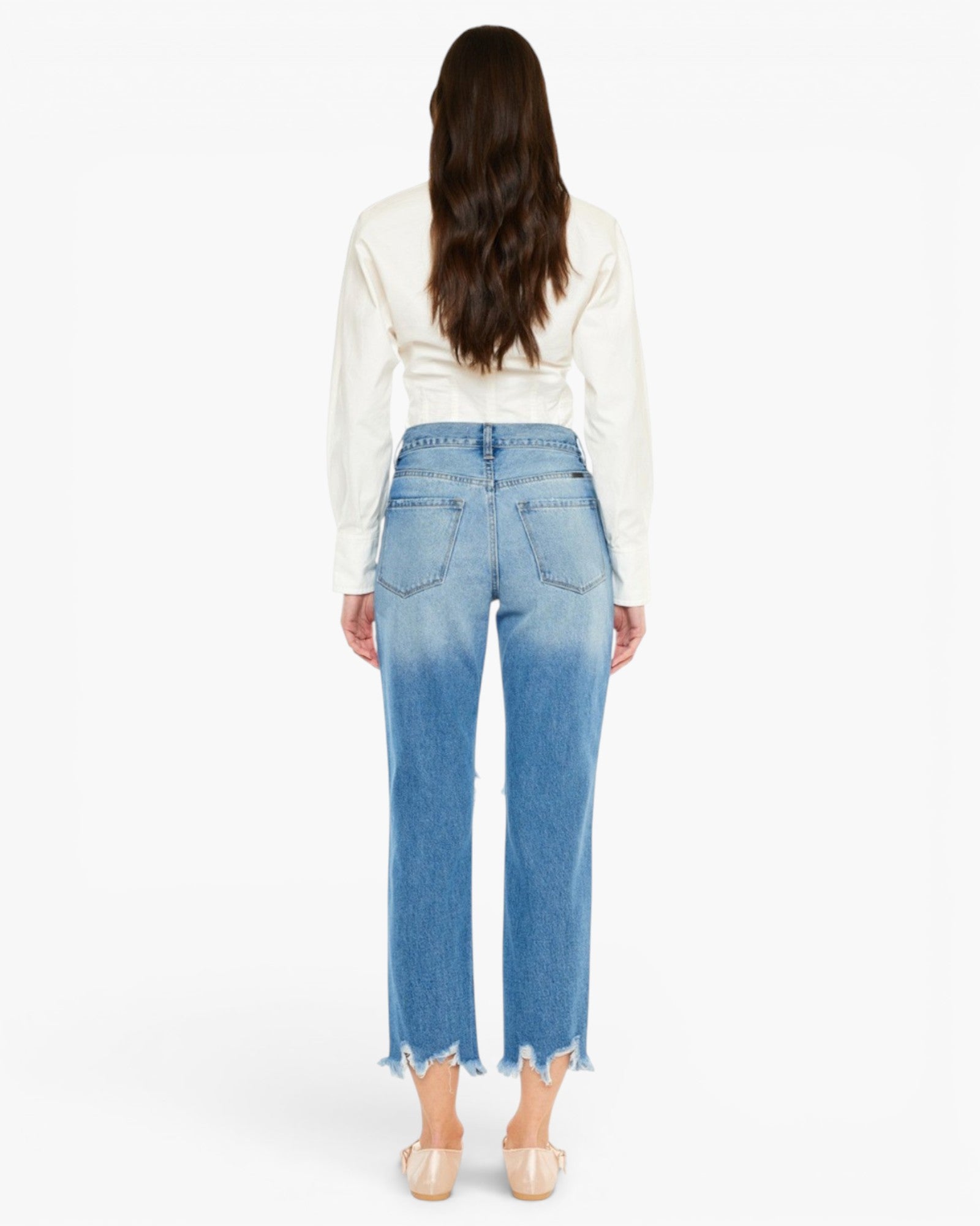 A woman viewed from the back wearing light blue cropped jeans with a faded wash, a cream long-sleeve top, and nude heels against a plain white studio background.